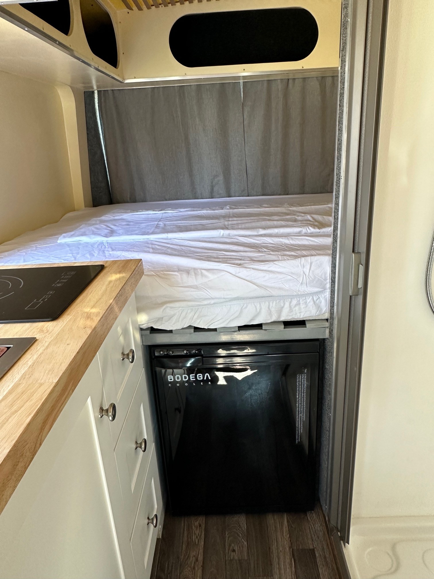 Compact RV rental interior featuring a cozy bed, kitchen counter with cooktop, and Bodega cooler.. FORD TRANSIT 2017