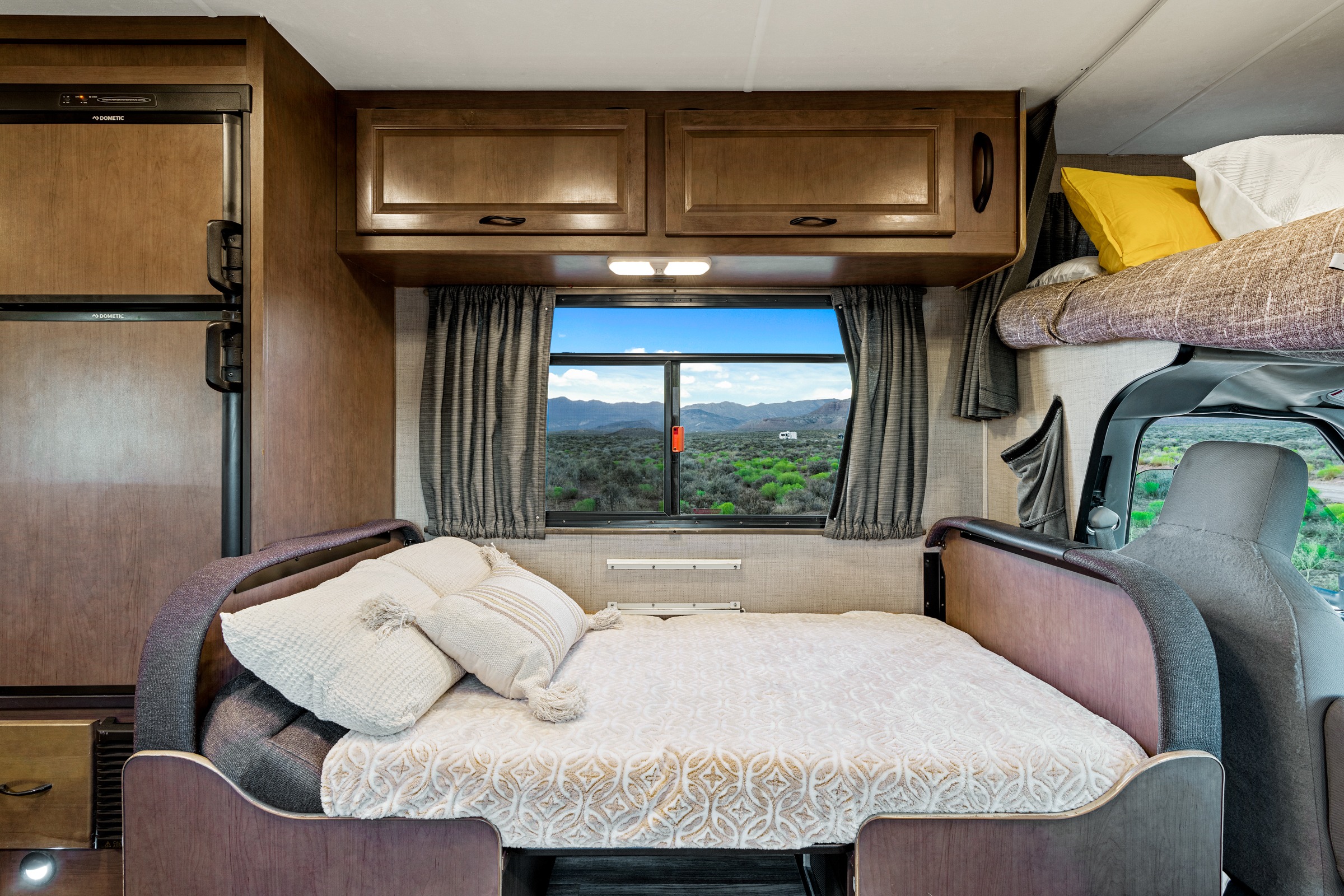 Comfortable RV rental interior featuring a main bed, overhead bunk, refrigerator, and a scenic desert window view.. Thor Majestic 2019