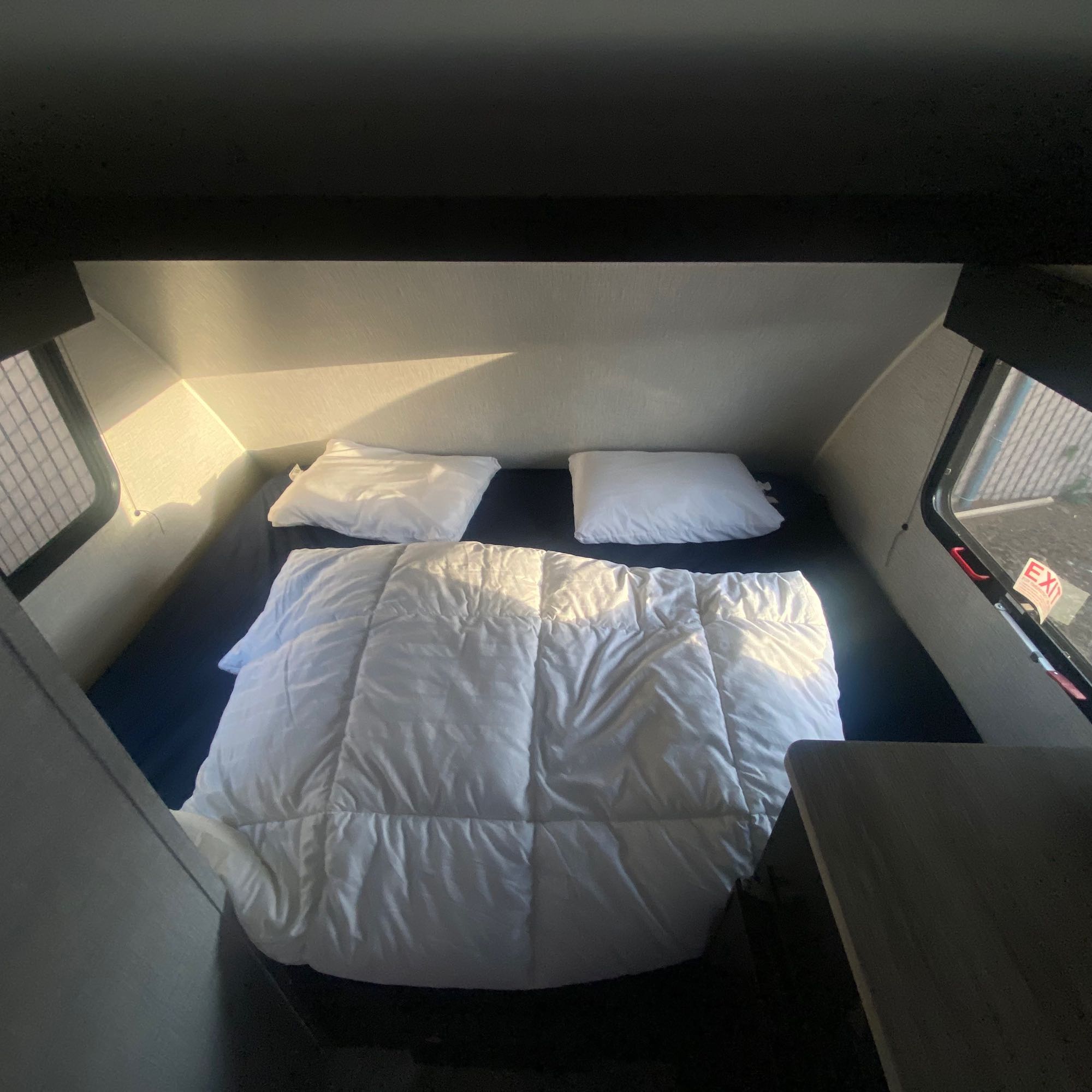 Cozy RV rental sleeping area with a bed, pillows, and white comforter, lit by sunlight from the window.. Braxton Creek Free Solo 2021