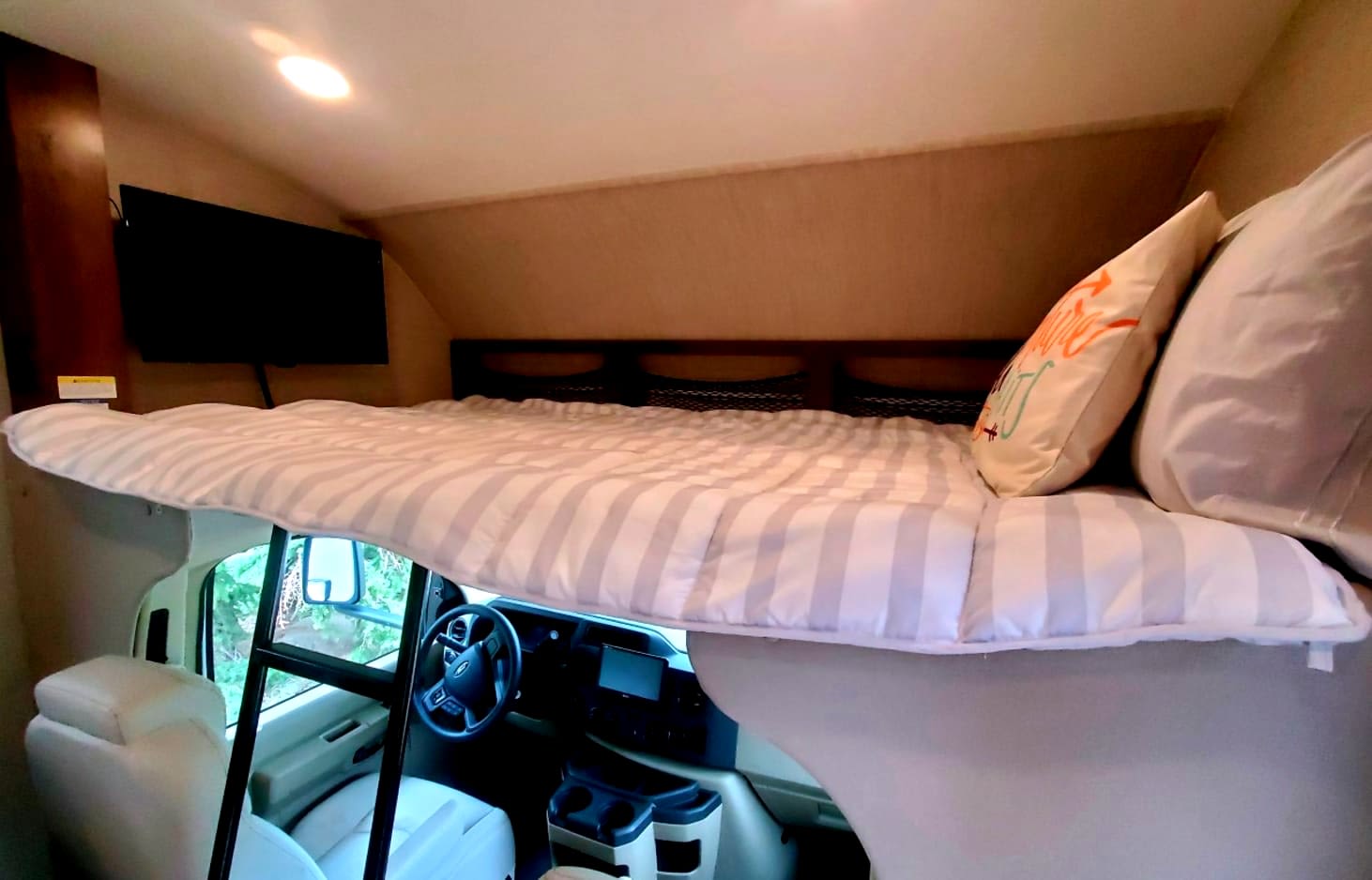 RV rental interior featuring a spacious overhead bunk with striped bedding, TV, and a view of the cab dashboard.. Jayco Redhawk 2022