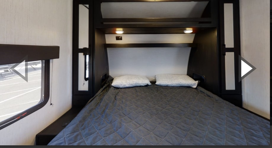 Comfortable RV rental bedroom with a large bed, dark comforter, pillows, overhead shelf, and reading lights, plus a window.. Heartland Fuel F-265 2022
