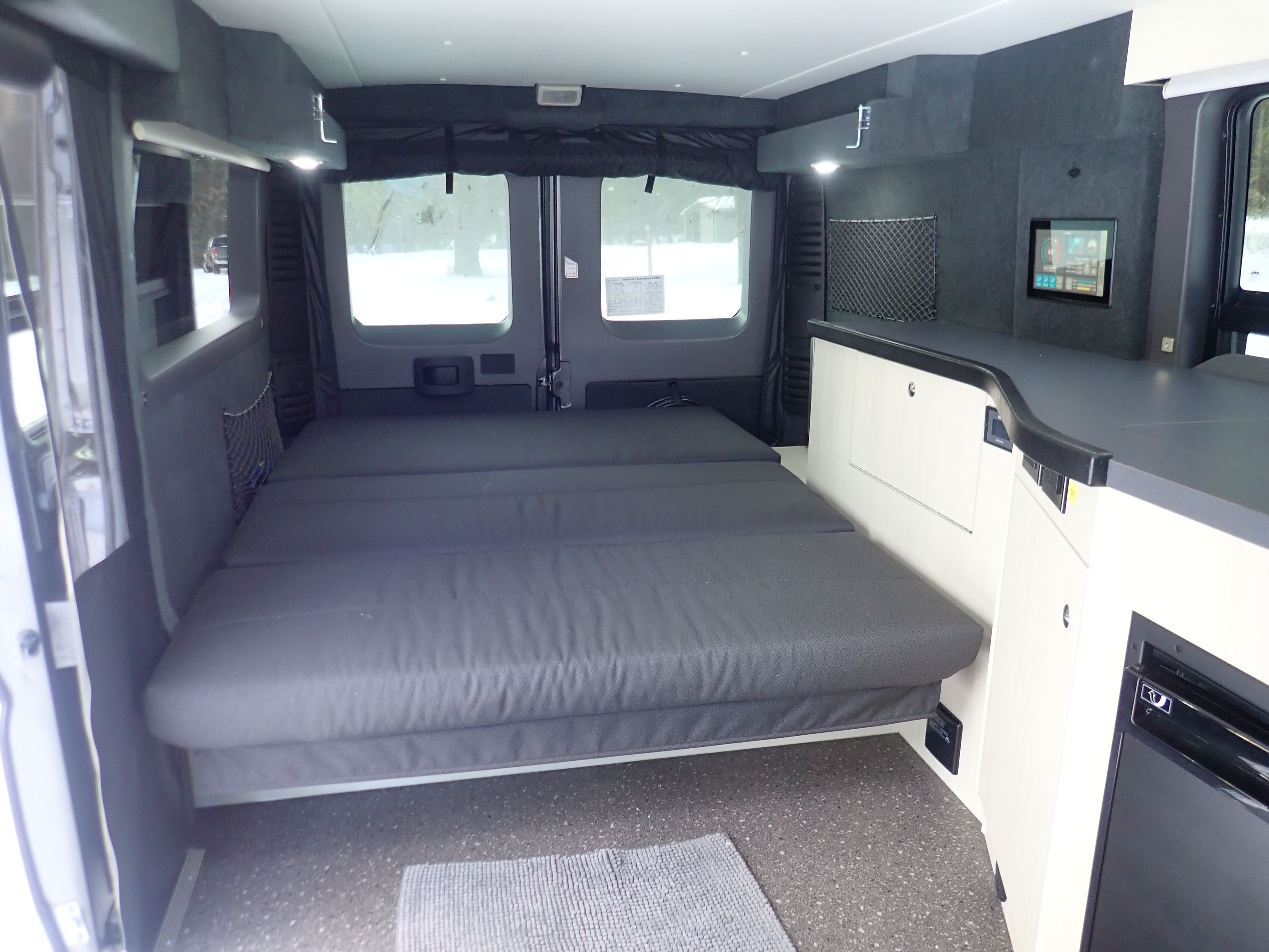 RV rental camper van interior showing a large grey bed, light walls, dark counters, and a wall-mounted screen.. Pleasure-Way Pleasure-Way Class B 2022