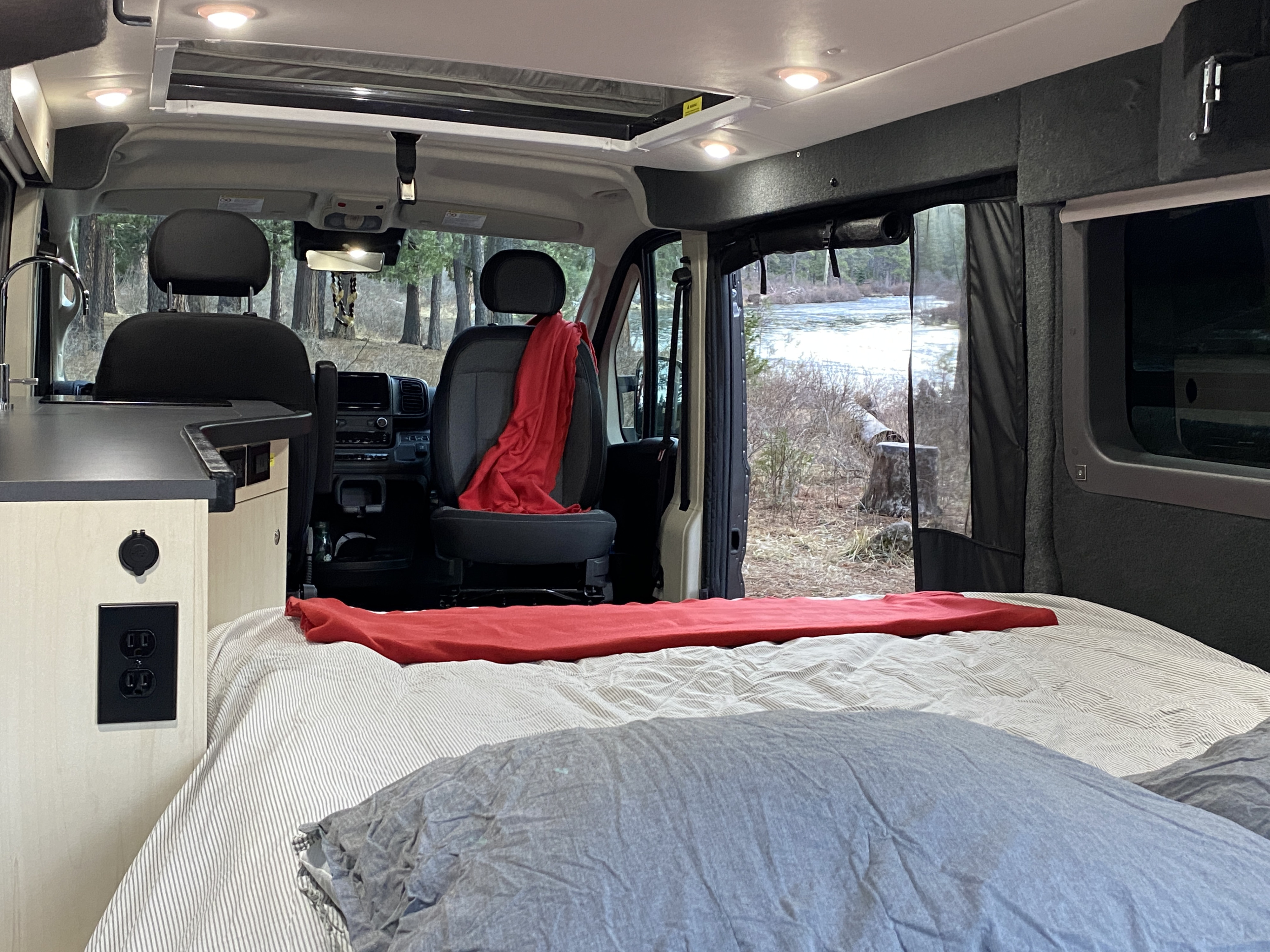 Cozy RV rental interior with a comfortable bed, kitchenette, and a peaceful view of a wintry river and trees outside.. Pleasure-Way Pleasure-Way Class B 2022