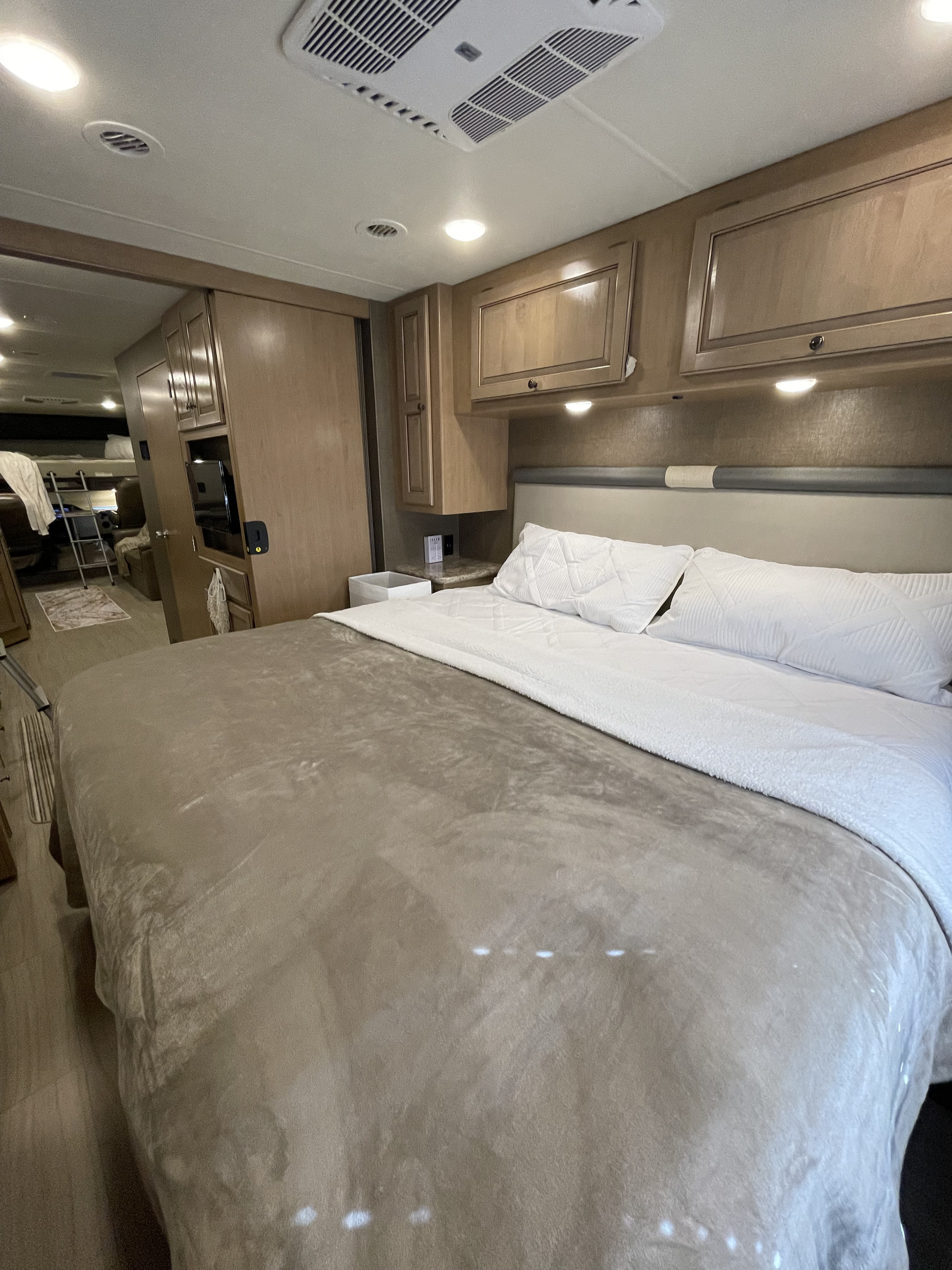 Cozy RV rental bedroom featuring a plush bed, overhead cabinets, and warm lighting. Hallway leads to bunks and entertainment.. Thor Windsport 2019