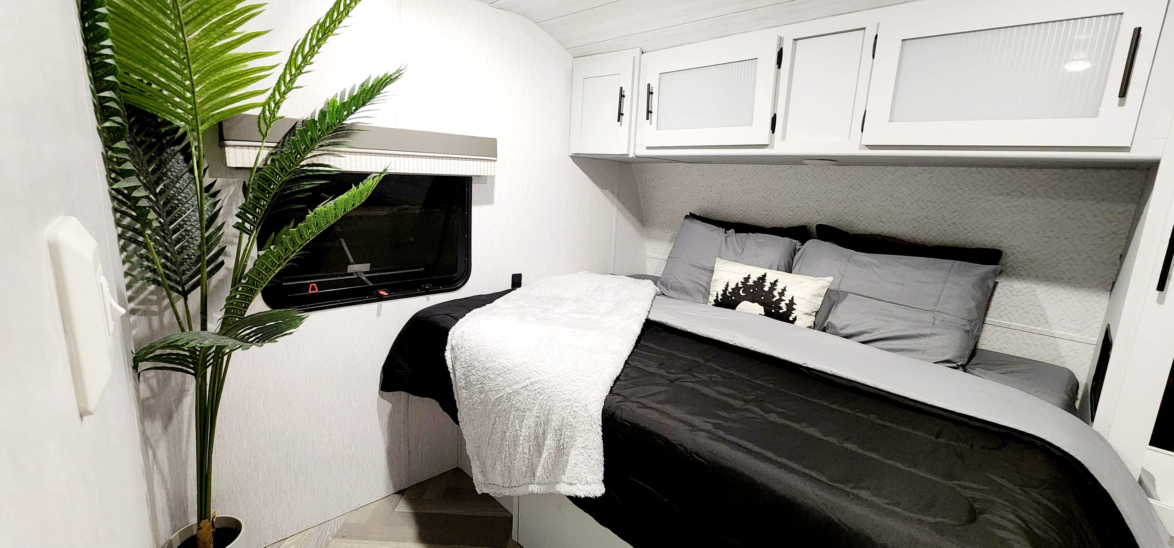 Cozy RV rental bedroom featuring a comfy bed with black & gray linens, white overhead cabinets, & a decorative plant.. Forest River Wildwood 2022