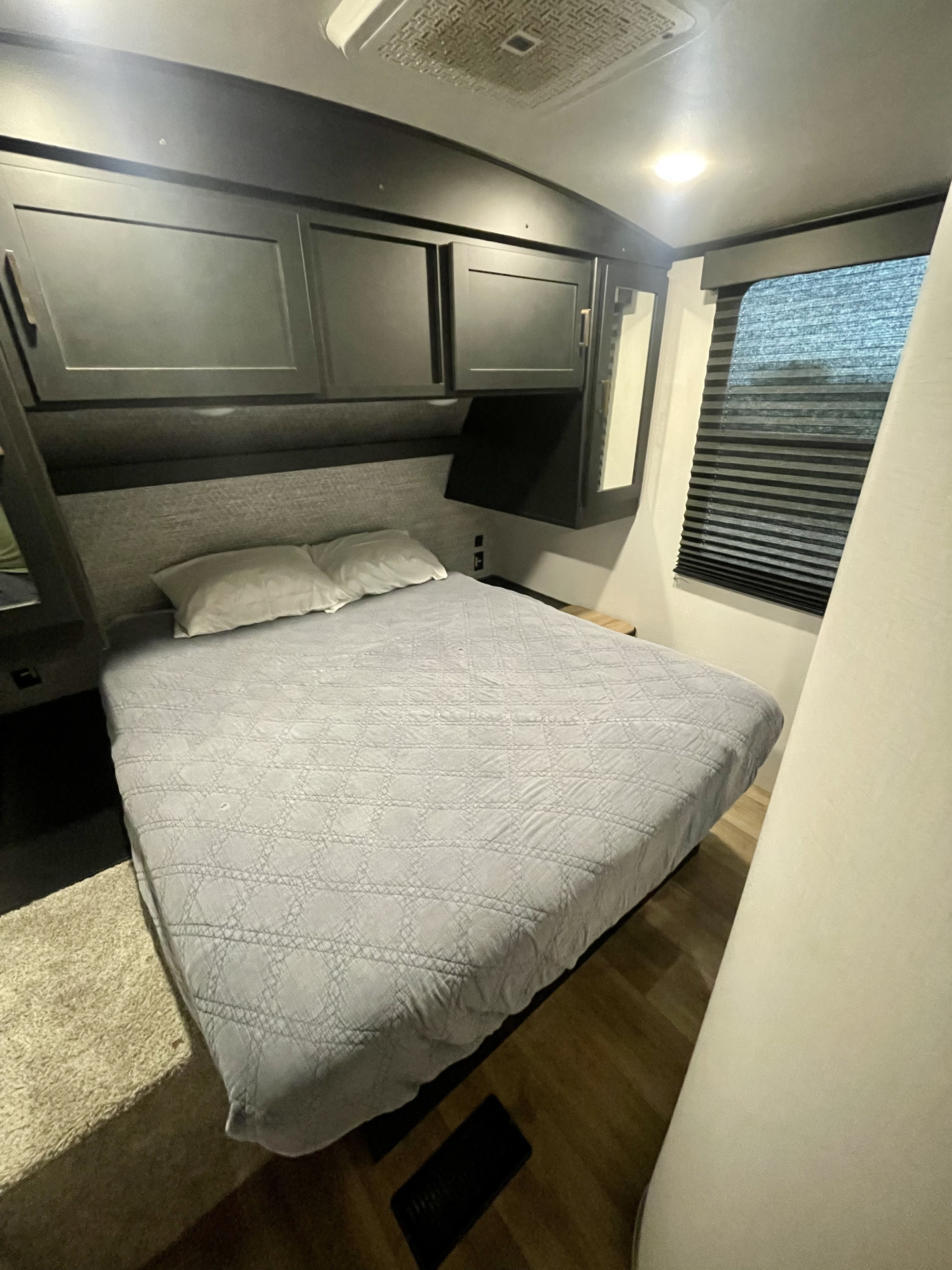 Inside an RV rental: a neat bedroom with a patterned bed, dark overhead cabinets, and window blinds, lit by ceiling lights.. Keystone Outback 2022