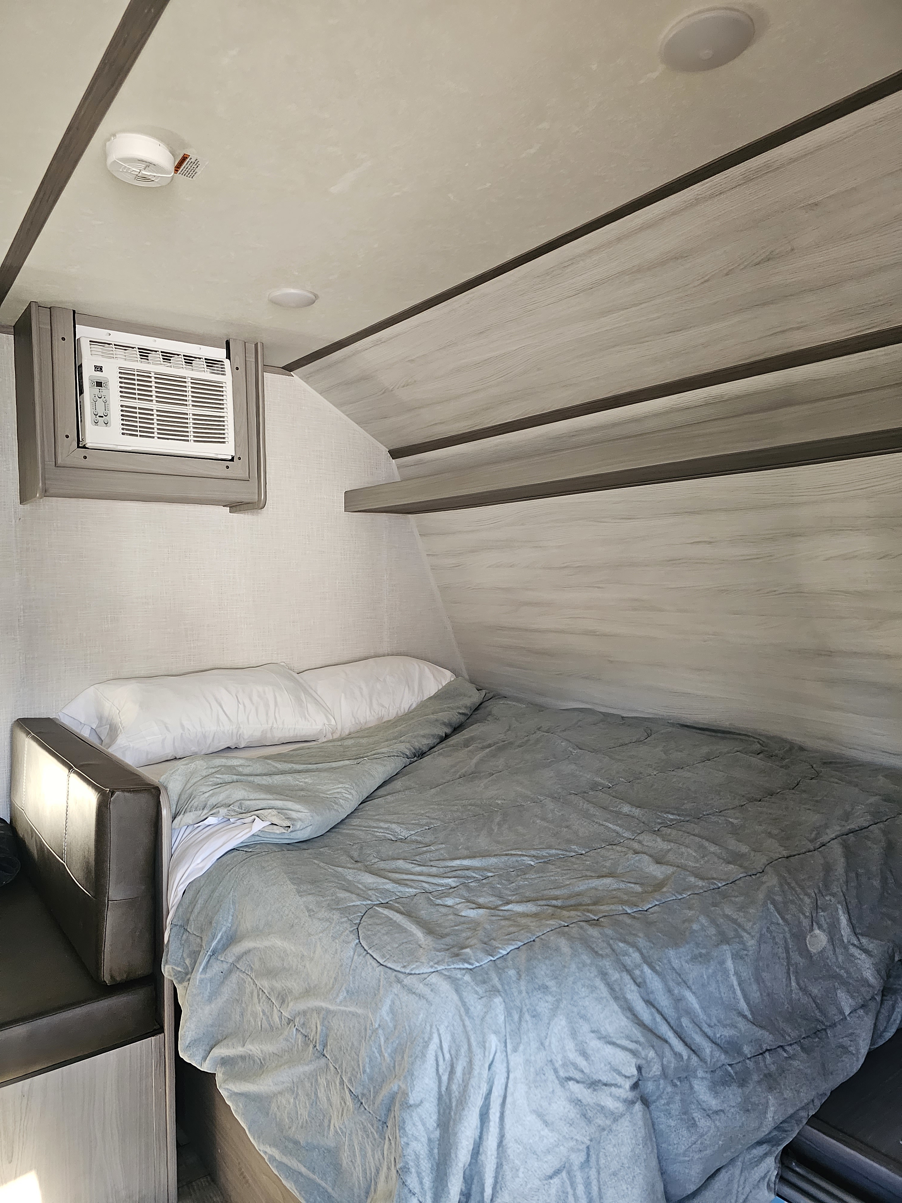 Compact RV rental bedroom showing a comfortable bed, window AC unit, and built-in shelving.. Dutchmen Coleman 17B 2023