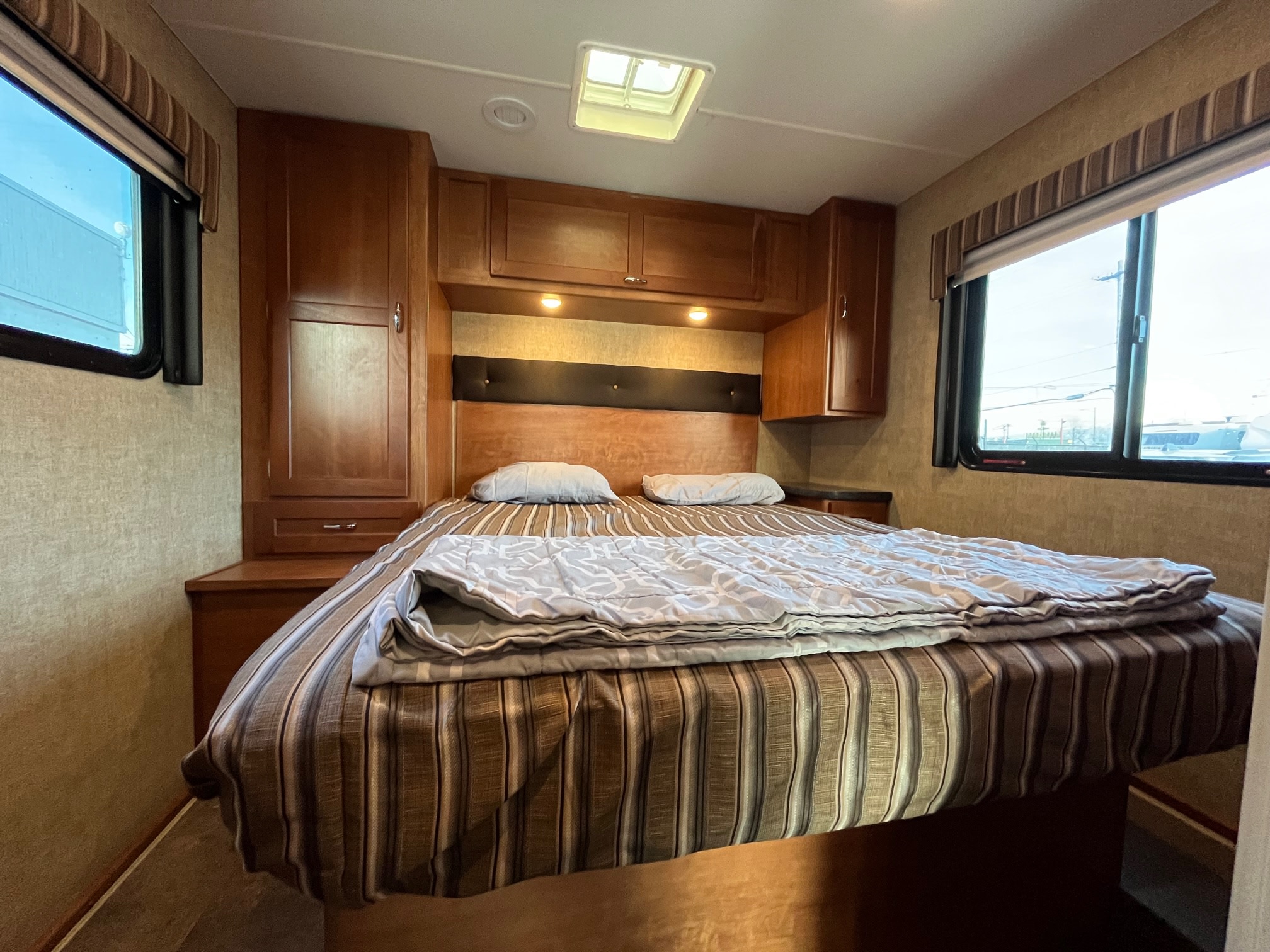 A comfortable RV rental bedroom features a large bed with striped bedding, wood cabinets, and bright windows, perfect for a trip.. Winnebago Minnie Winnie 2017