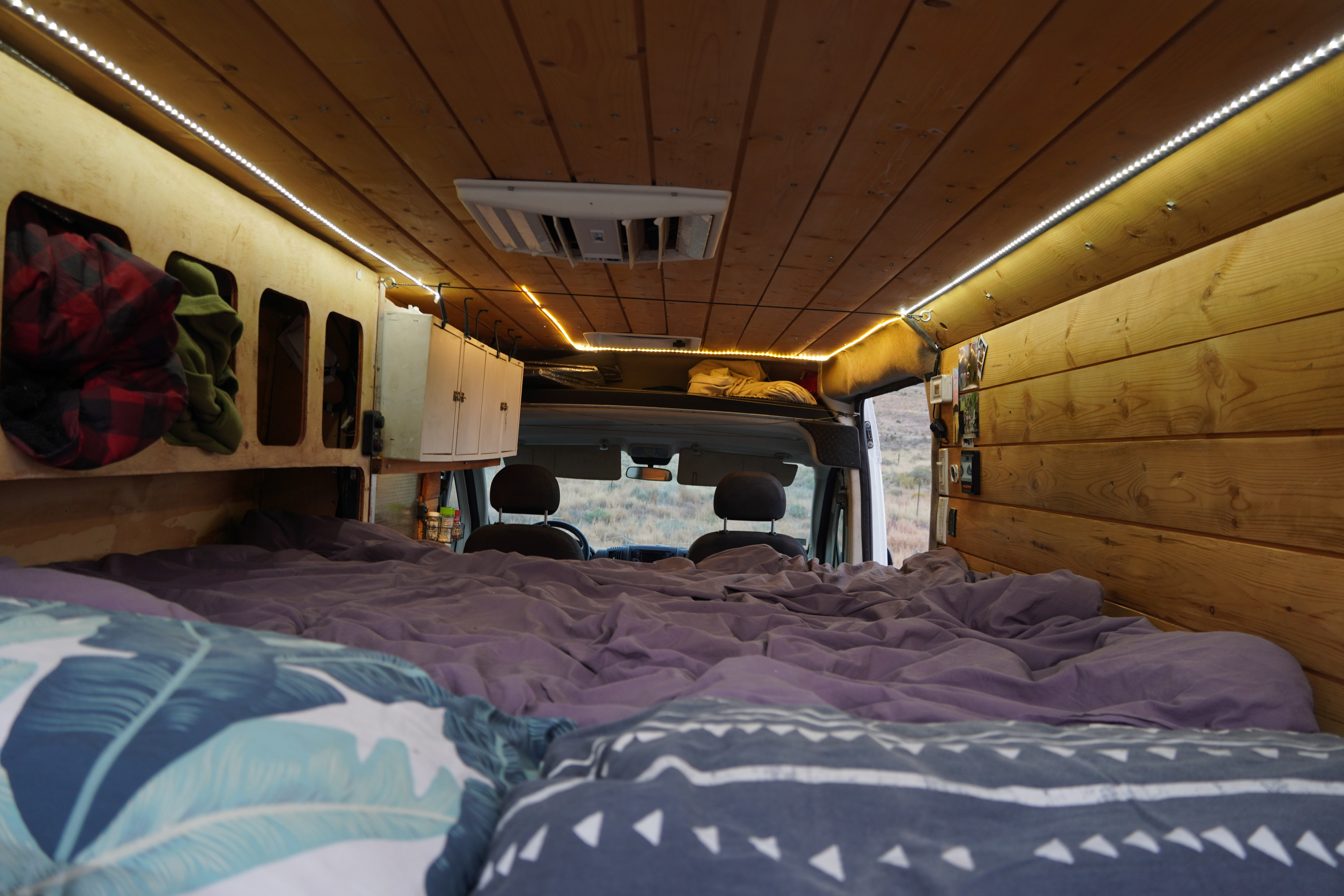 An RV rental's cozy interior: large bed, wood paneling, LED lights, and cab view of a dry landscape.. Dodge Promaster 2500 2017