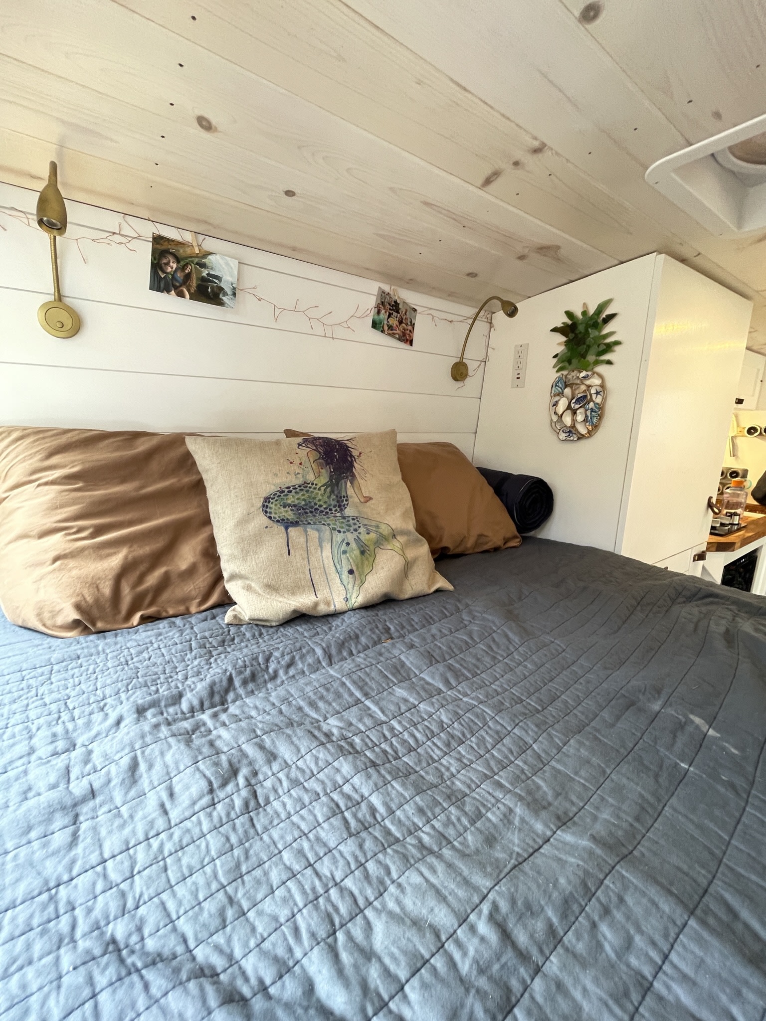 Cozy RV rental bed area with a mermaid pillow, soft quilt, wall lamps, and string lights with photos above.. Ford Transit 250 2019