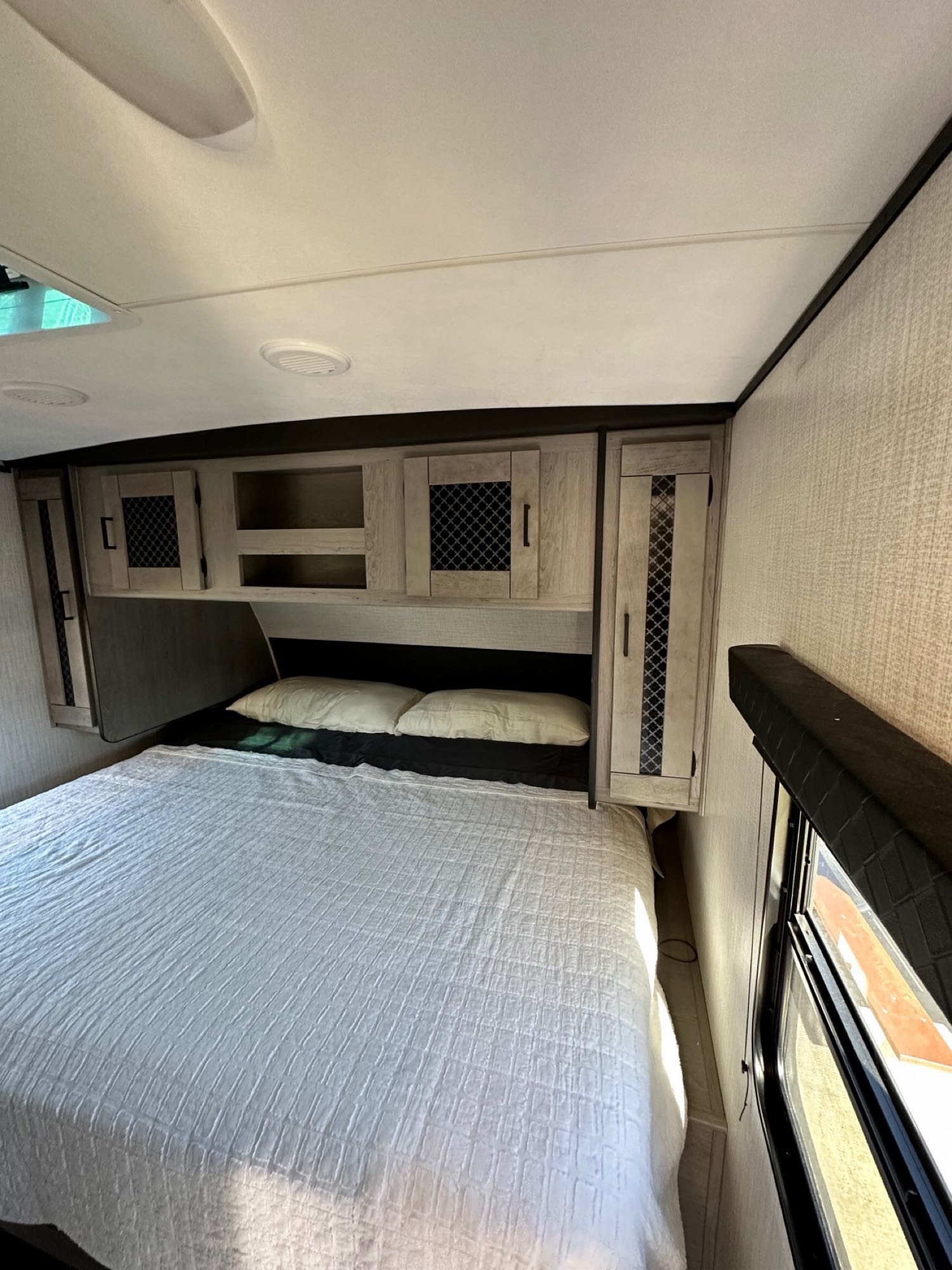 RV rental bedroom with a bed, white bedding, overhead cabinets, and a window, offering a cozy sleeping space.. Heartland RVs Mallard 2022