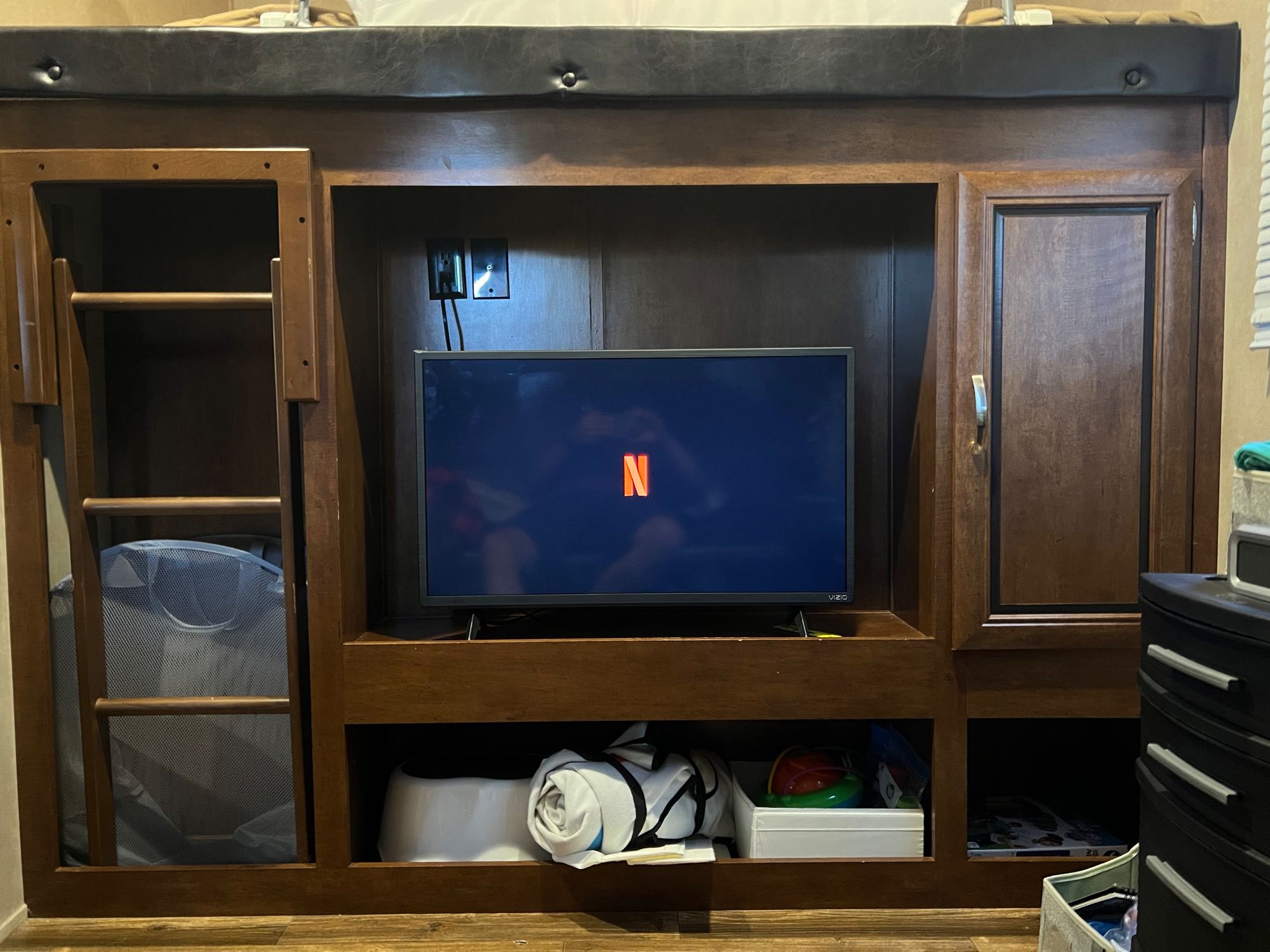 Inside an RV rental: a wooden entertainment center with a TV showing Netflix, storage cubbies, and a bunk bed ladder.. Forest River Vibe 2017