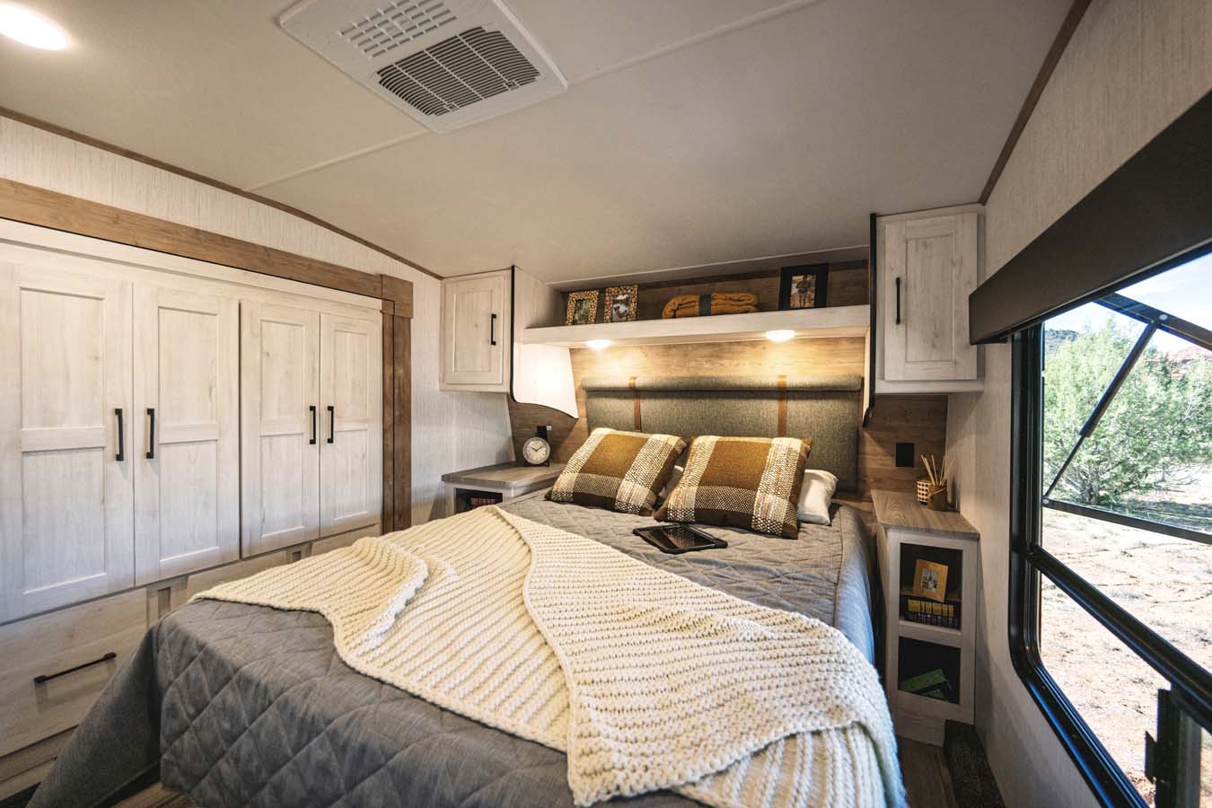Modern RV rental bedroom featuring a comfortable bed, overhead storage, wardrobe, and a large window overlooking nature.. Heartland RVs Milestone 2023