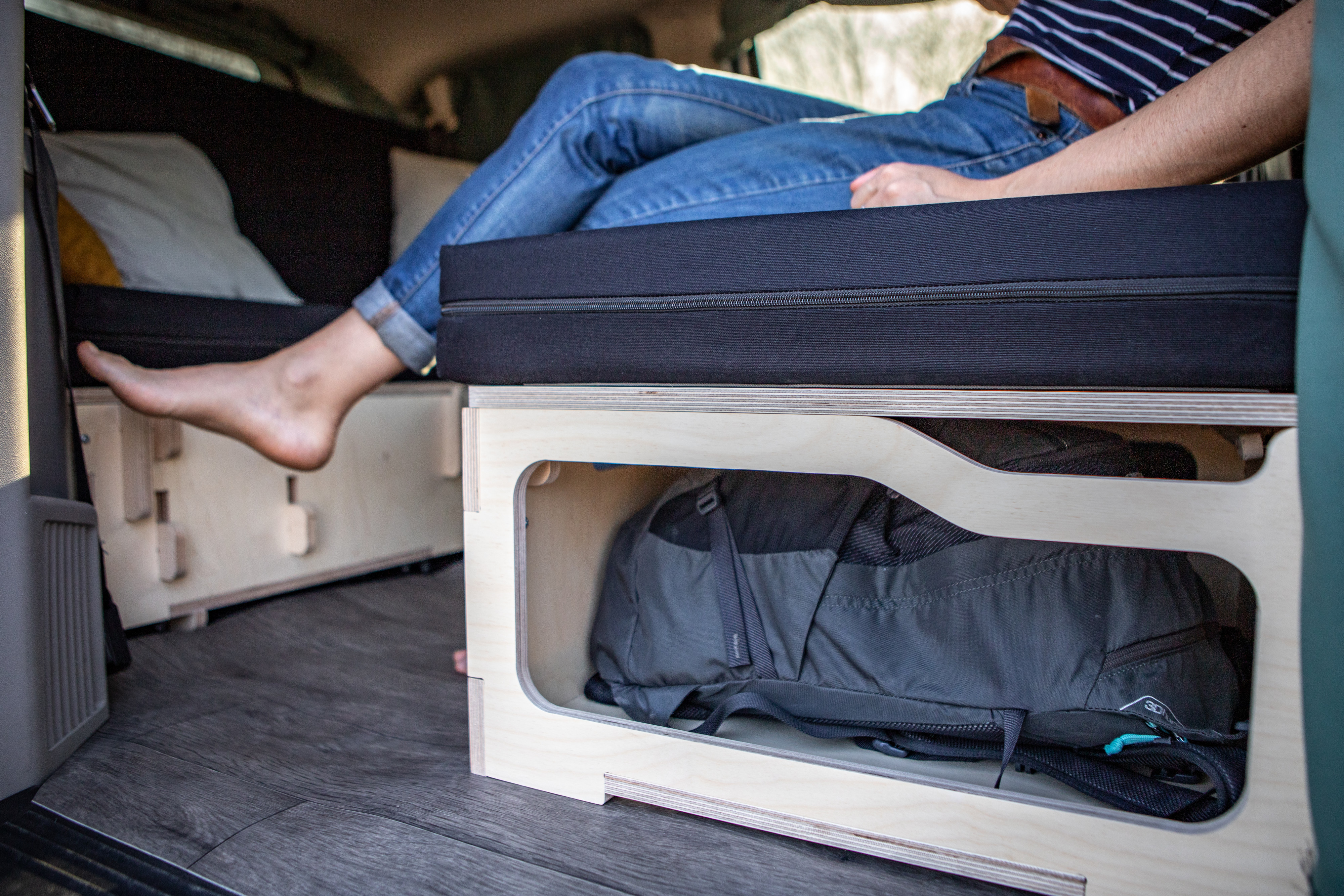 Enjoy your rv rental's functional interior. A person relaxes on a cushioned bed, with clever wooden storage below for a backpack.. Chrysler Grand Caravan 2025