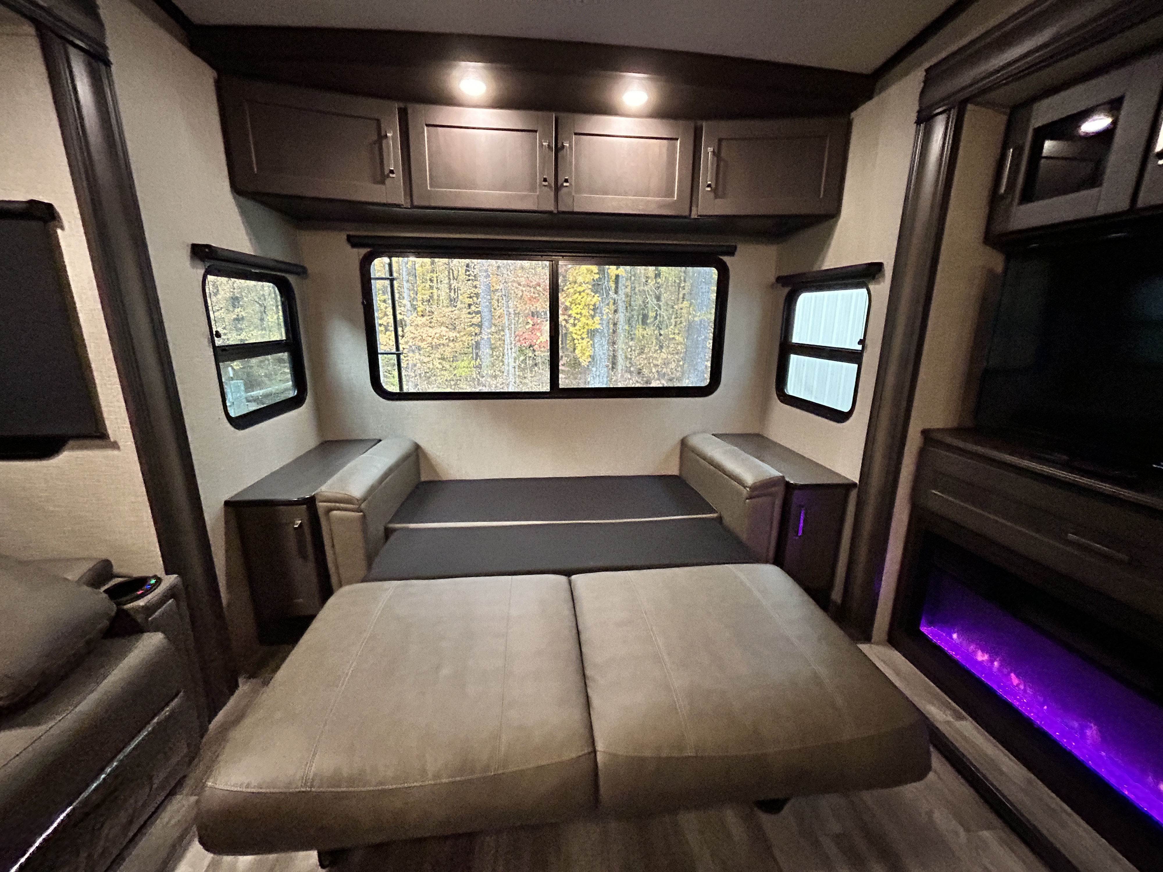 Spacious RV rental living area featuring a sofa bed, bright windows with forest views, cabinets, and a glowing fireplace.. Grand Design Reflection 2022