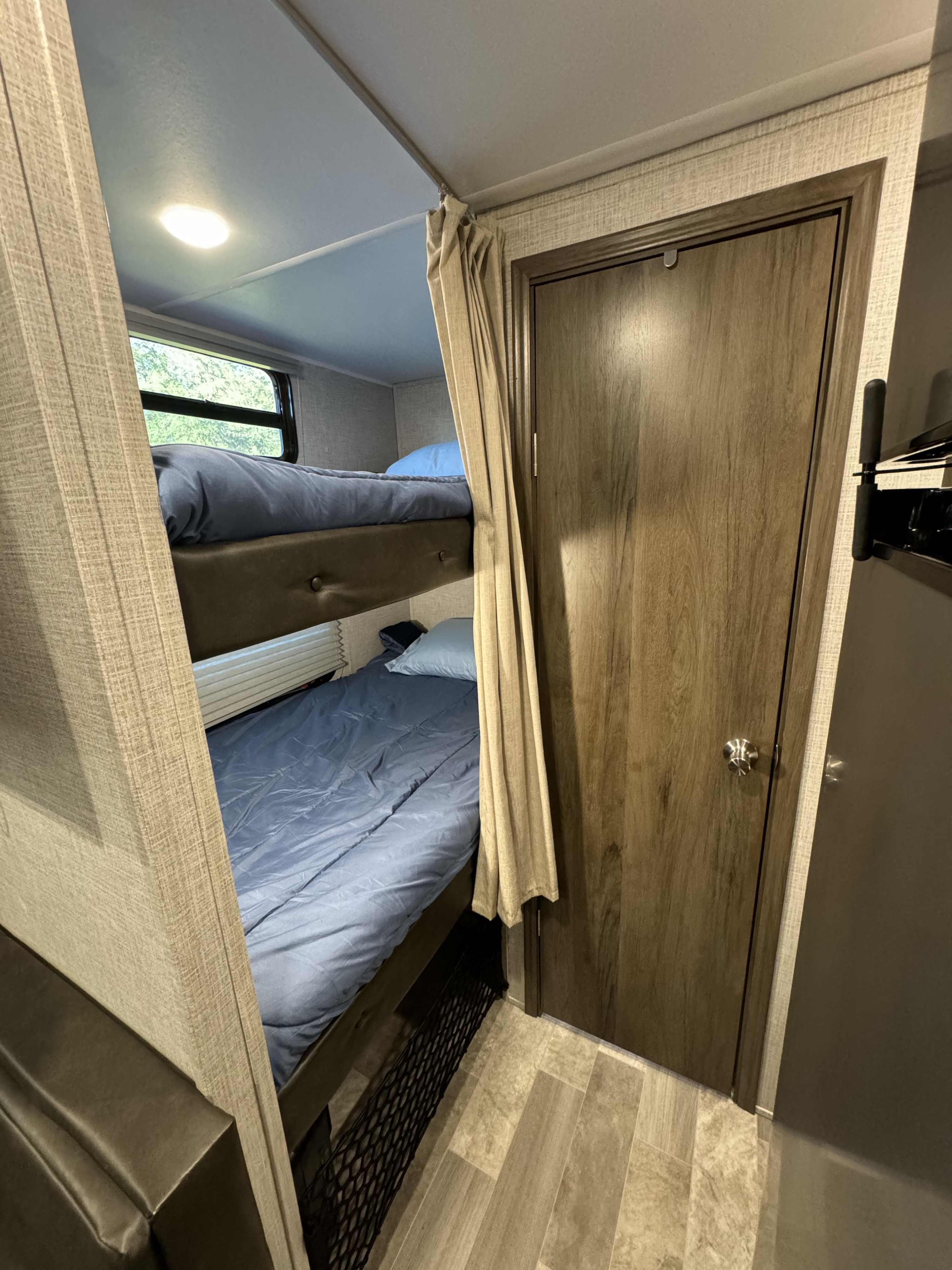 RV rental interior featuring cozy bunk beds with blue bedding, a window view, privacy curtain, and wooden door.. East to West Della Terra 2021