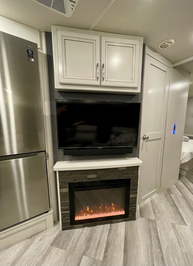 Modern RV rental interior: stainless fridge, TV, electric fireplace with stone facade, & light cabinets. Bedroom visible.. Fleetwood Bounder 33C 2022
