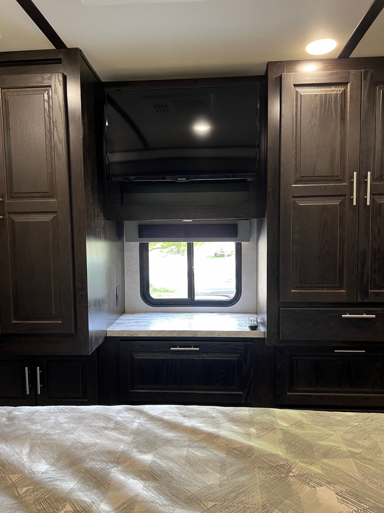 RV rental bedroom with dark wood built-in cabinetry, TV, and window. A patterned bedspread fills the foreground.. Forest River Fr3 2020