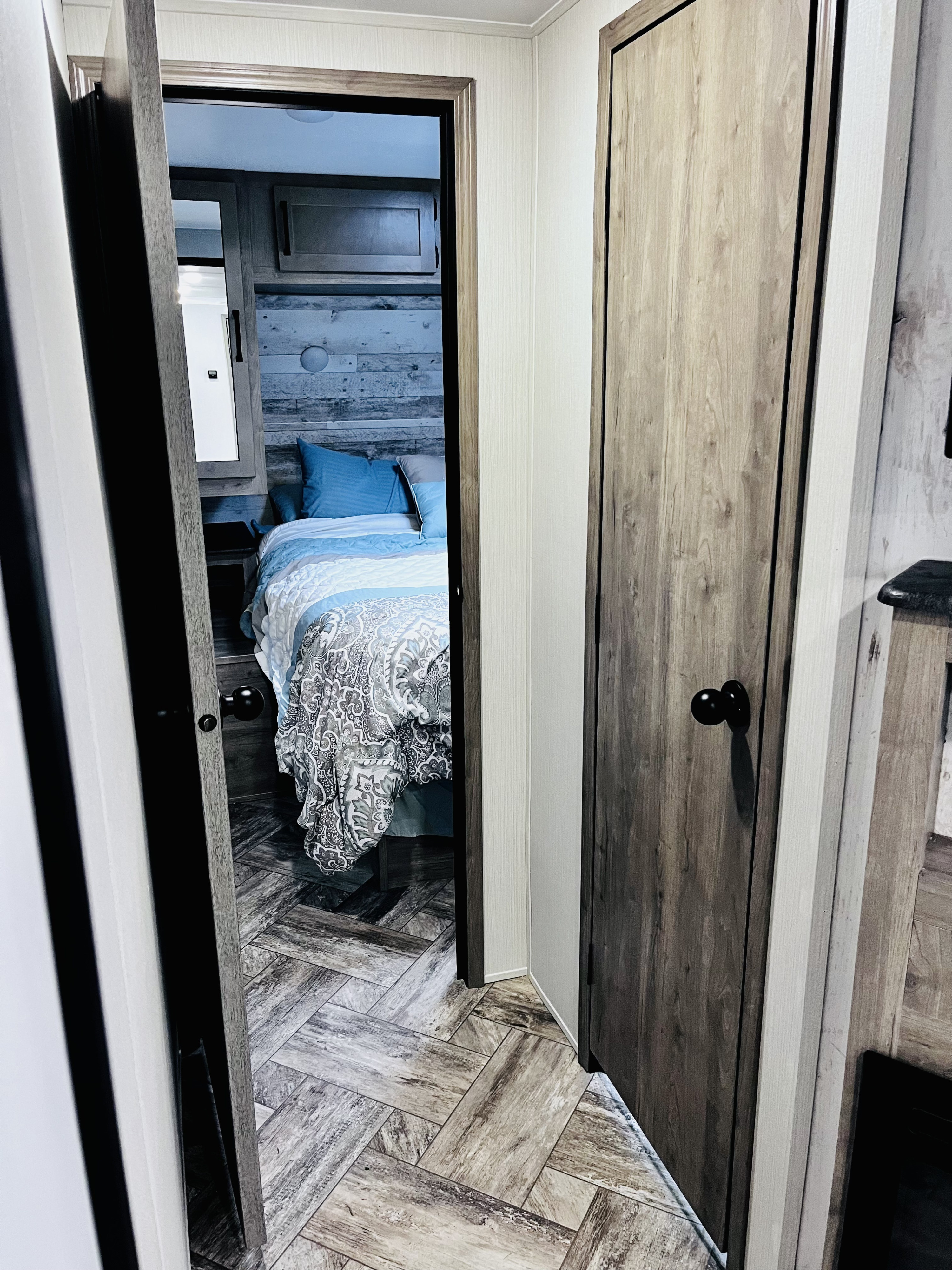 Looking down an RV rental hallway into a bright bedroom with blue and patterned bedding, a wood headboard, and herringbone floors.. Forest River Puma 28bhss 2024