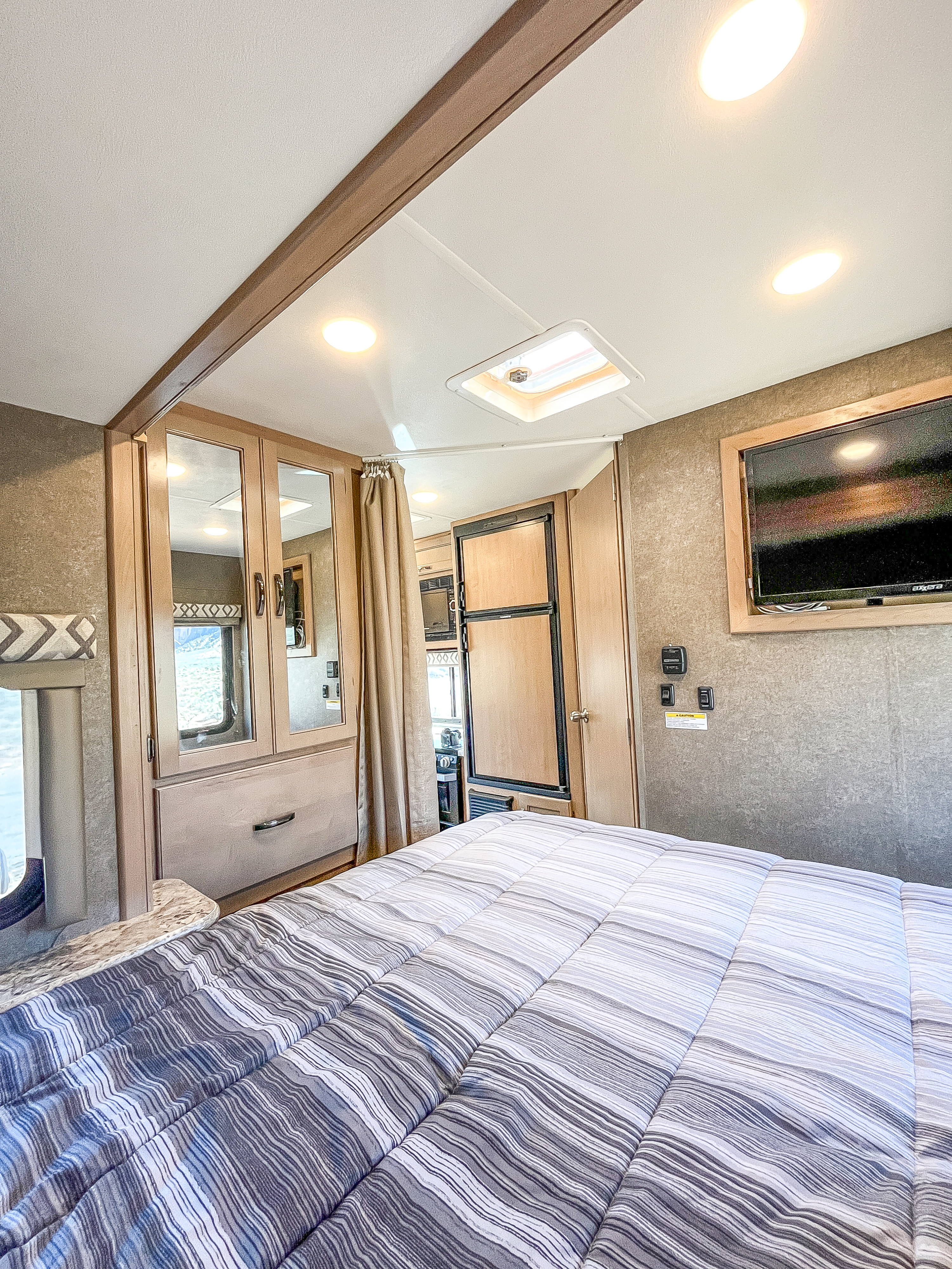 Spacious RV rental bedroom area with a striped bed, built-in storage, TV, and kitchenette appliances like a fridge and microwave.. Thor Four Winds 2019