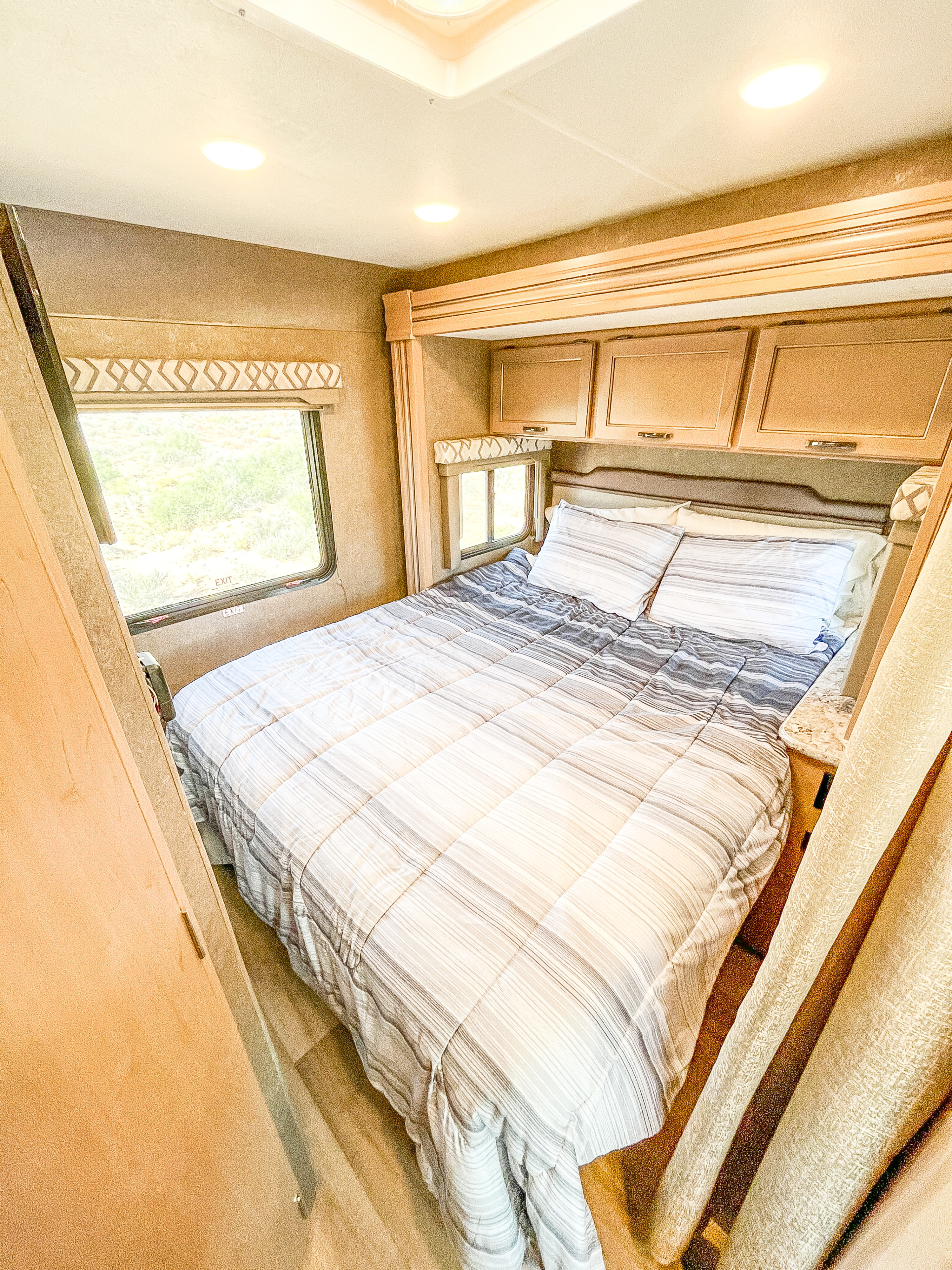 Cozy RV rental bedroom with striped bedding, overhead cabinets, and bright windows offering a comfortable interior.. Thor Four Winds 2019