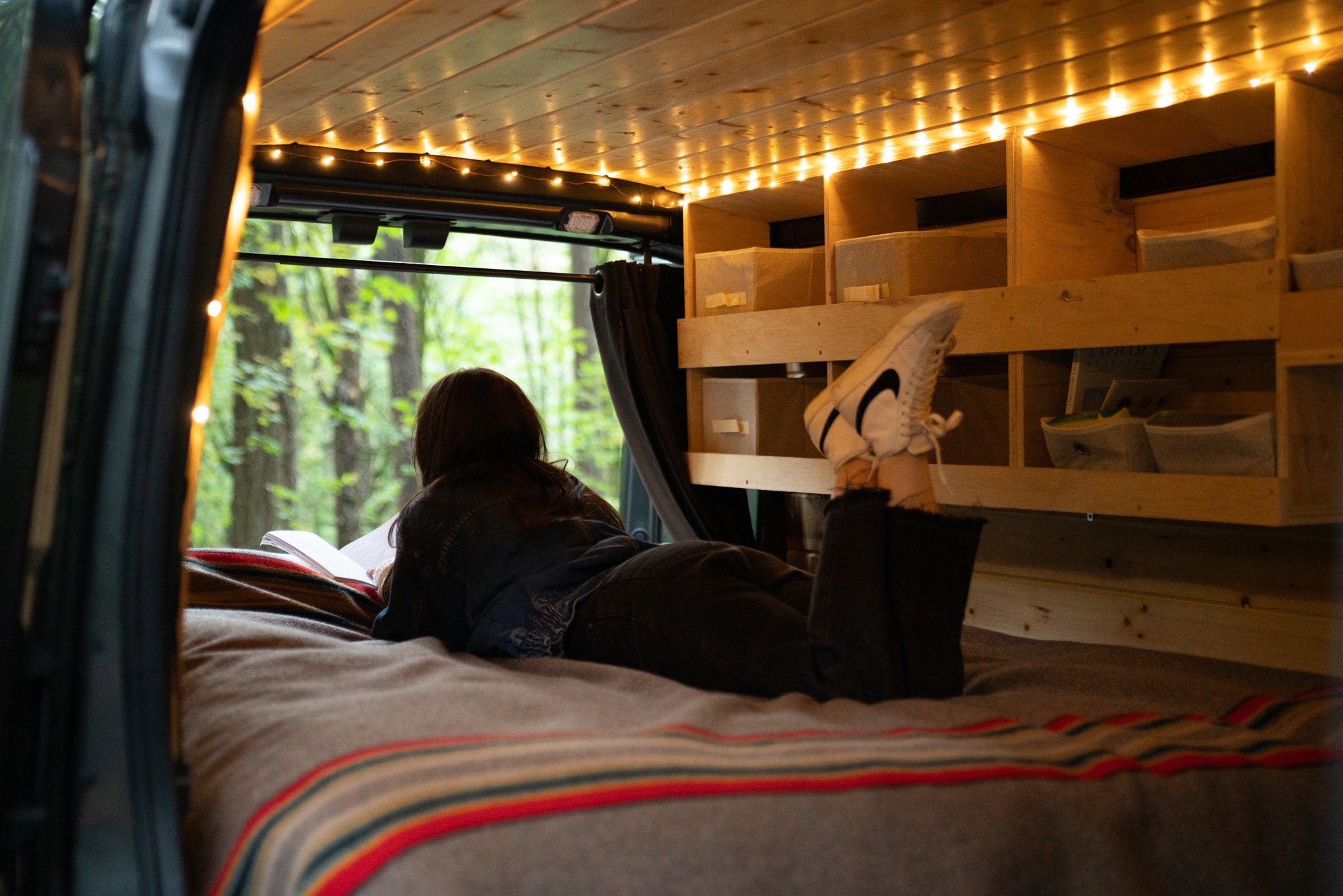 Relax in your **rv rental**! A person reads a book from a cozy bed inside a wooden camper van, with ambient lights & forest views.. Ford Transit 2020