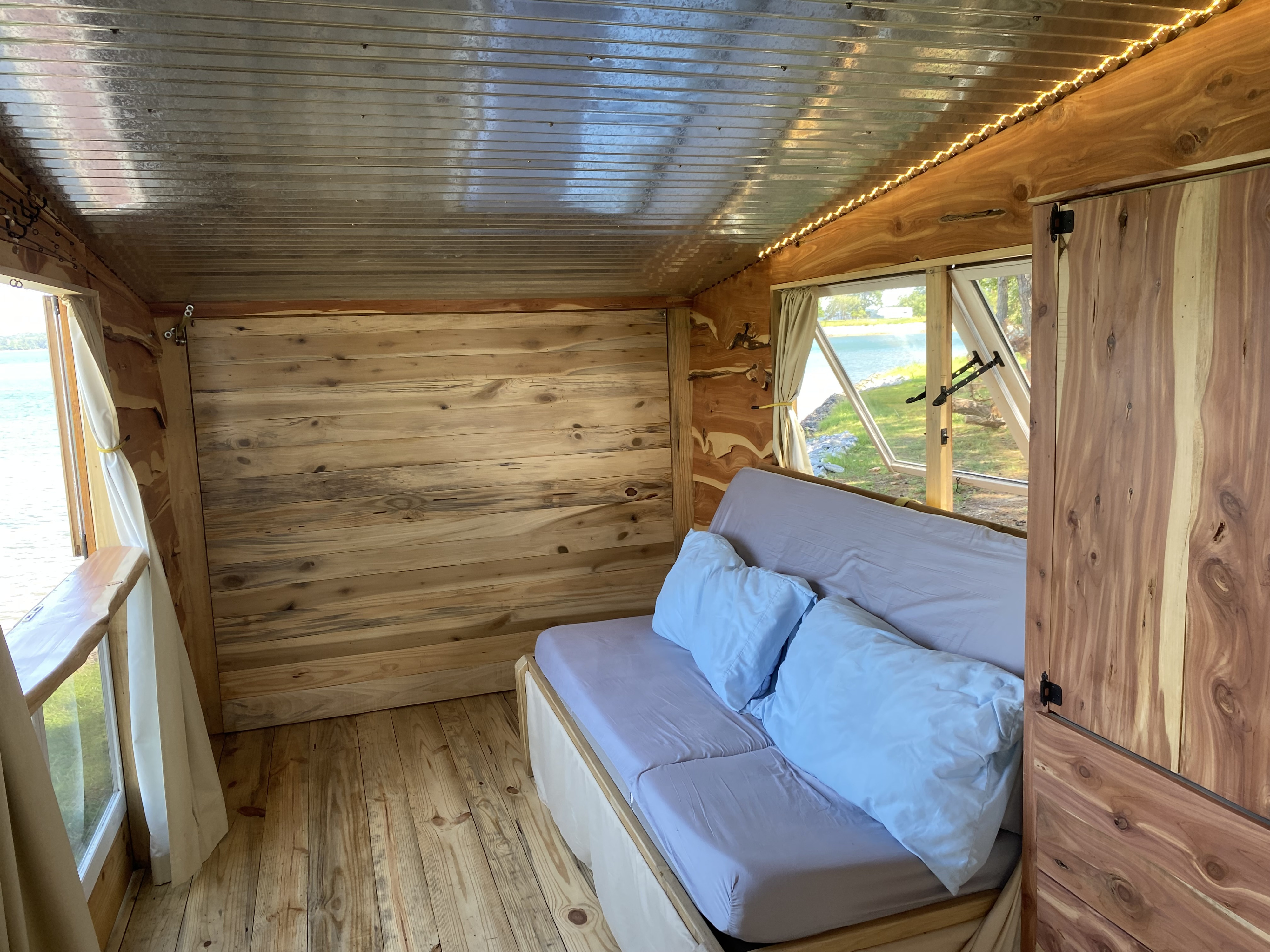 Explore this rustic RV rental interior with natural wood, cozy futon, and scenic lake views from large windows.. Tiny House On Wheels Glamper / Glamping Trailer THOW Tiny House On Wheels Glamper / Glamping Trailer THOW 2023