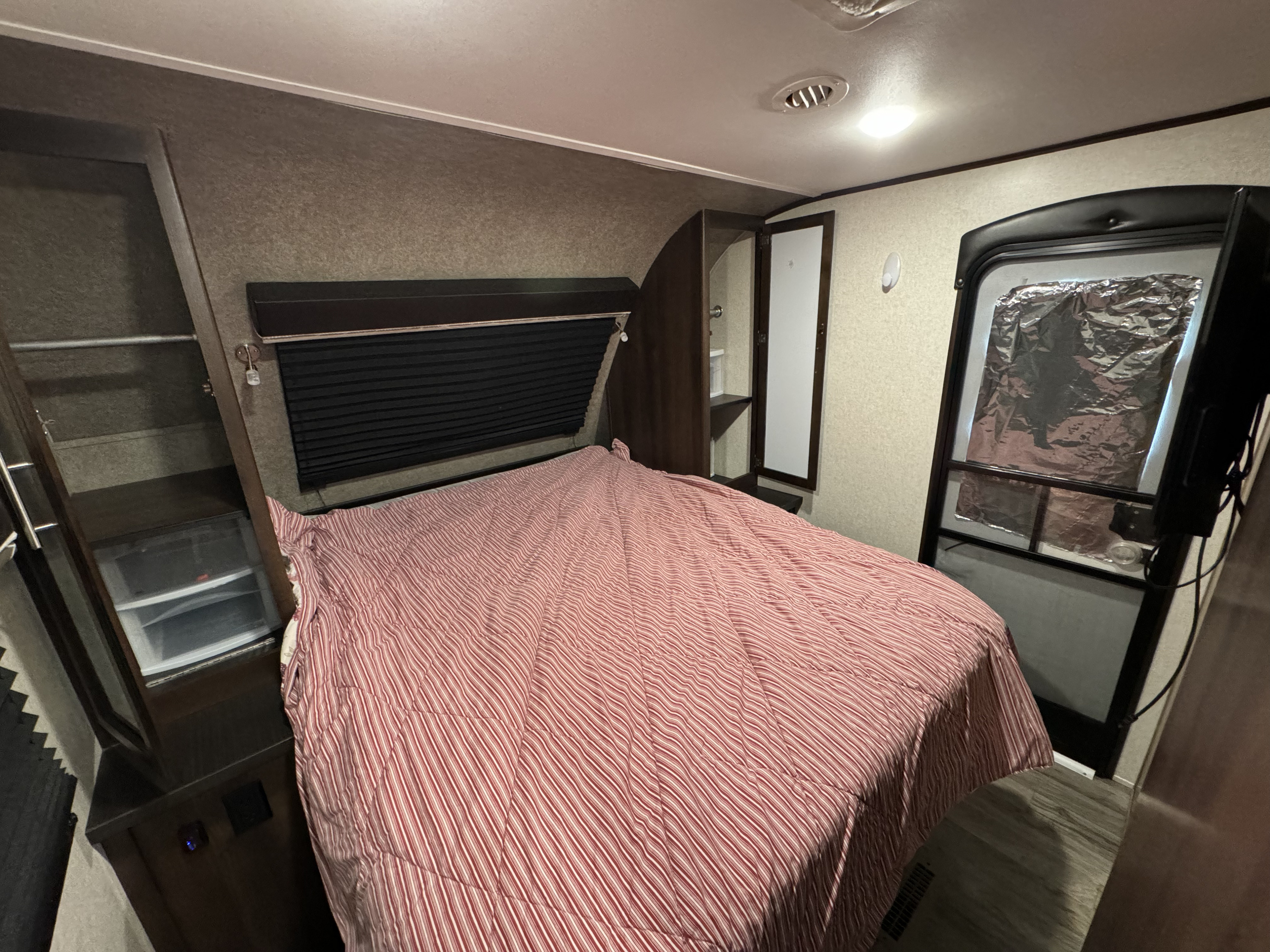 Cozy RV rental bedroom with a striped bed, built-in storage cabinets, and a privacy-covered window by the entry door.. Jayco White Hawk 2018