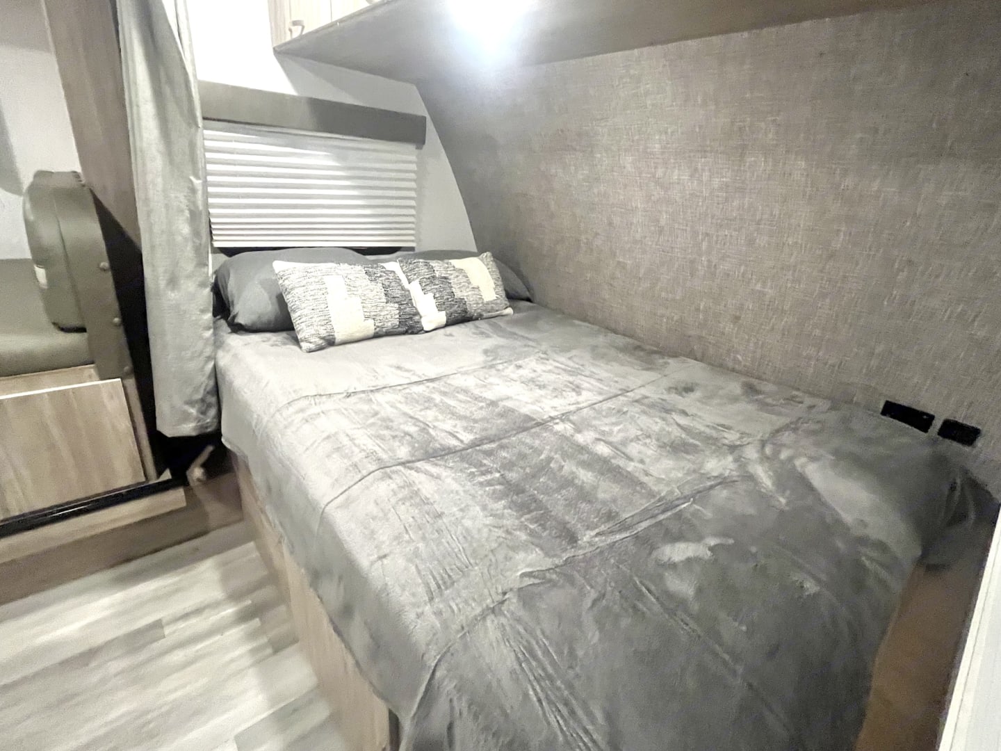 Cozy RV rental bedroom featuring a grey bed with patterned pillows, textured walls, and a window with blinds.. Winnebago Minnie Winnie 2022