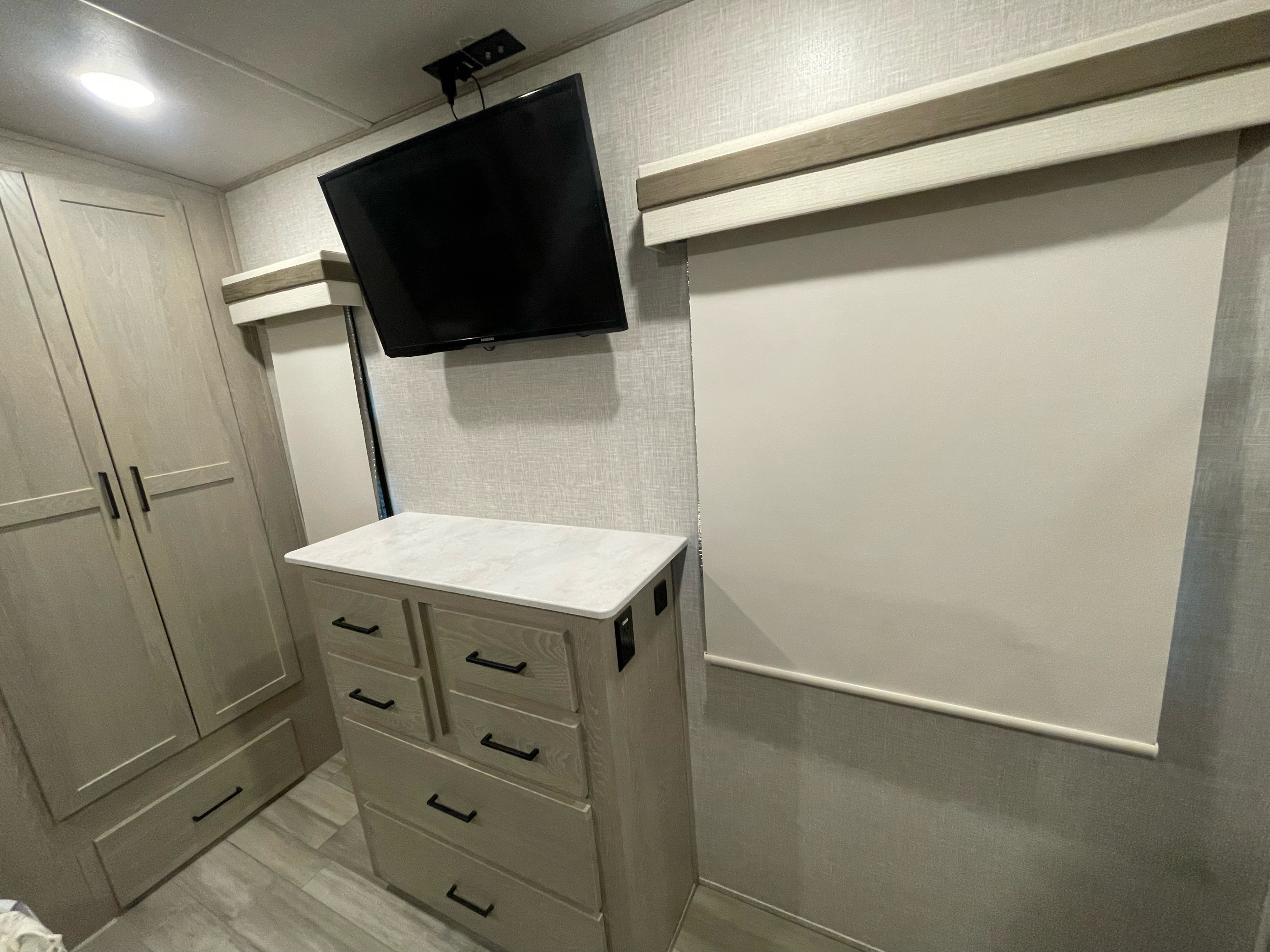 Modern RV rental interior: bedroom space with a wall-mounted TV, dresser, wardrobe, and a pulled-down window blind.. Forest River Rockwood Signature 2022