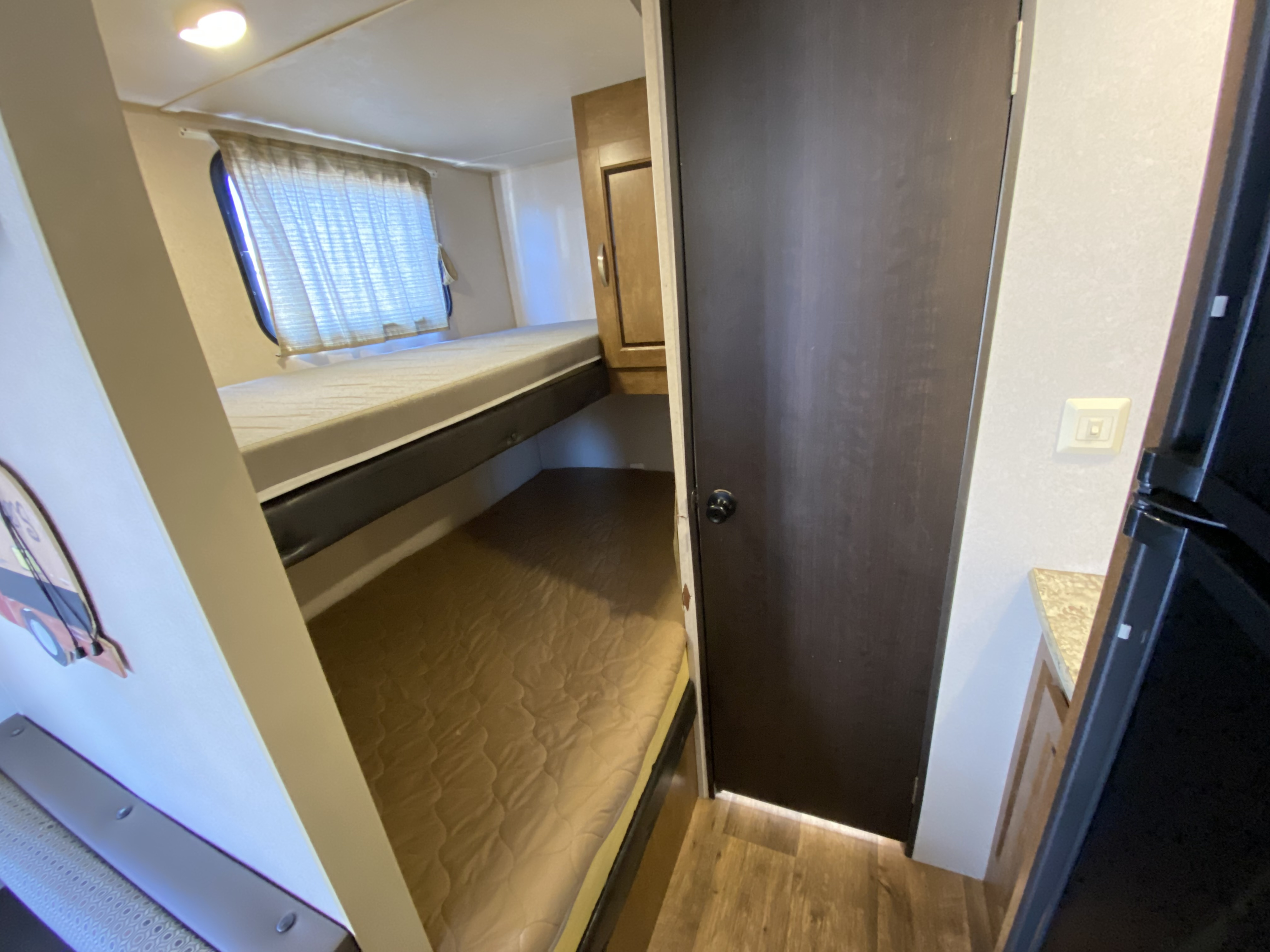 Inside an RV rental: bunk beds with mattresses, a window, a dark brown door, and partial view of a refrigerator.. Forest River Salem Cruise Lite 2017