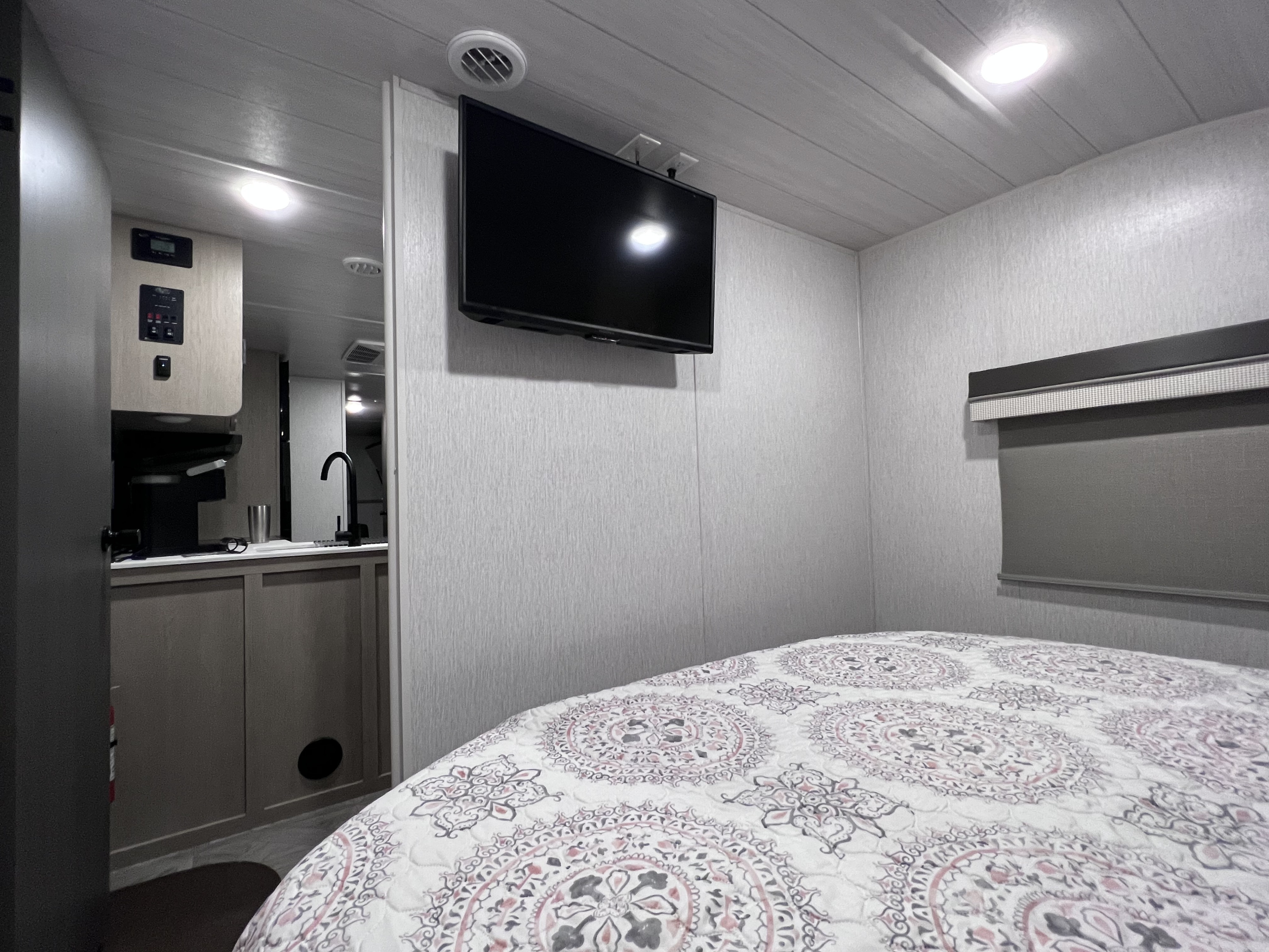 Inside an RV rental: cozy bedroom with patterned quilt, wall-mounted TV, and a compact kitchen area.. Forest River Wildwood X-Lite Platinum 2023