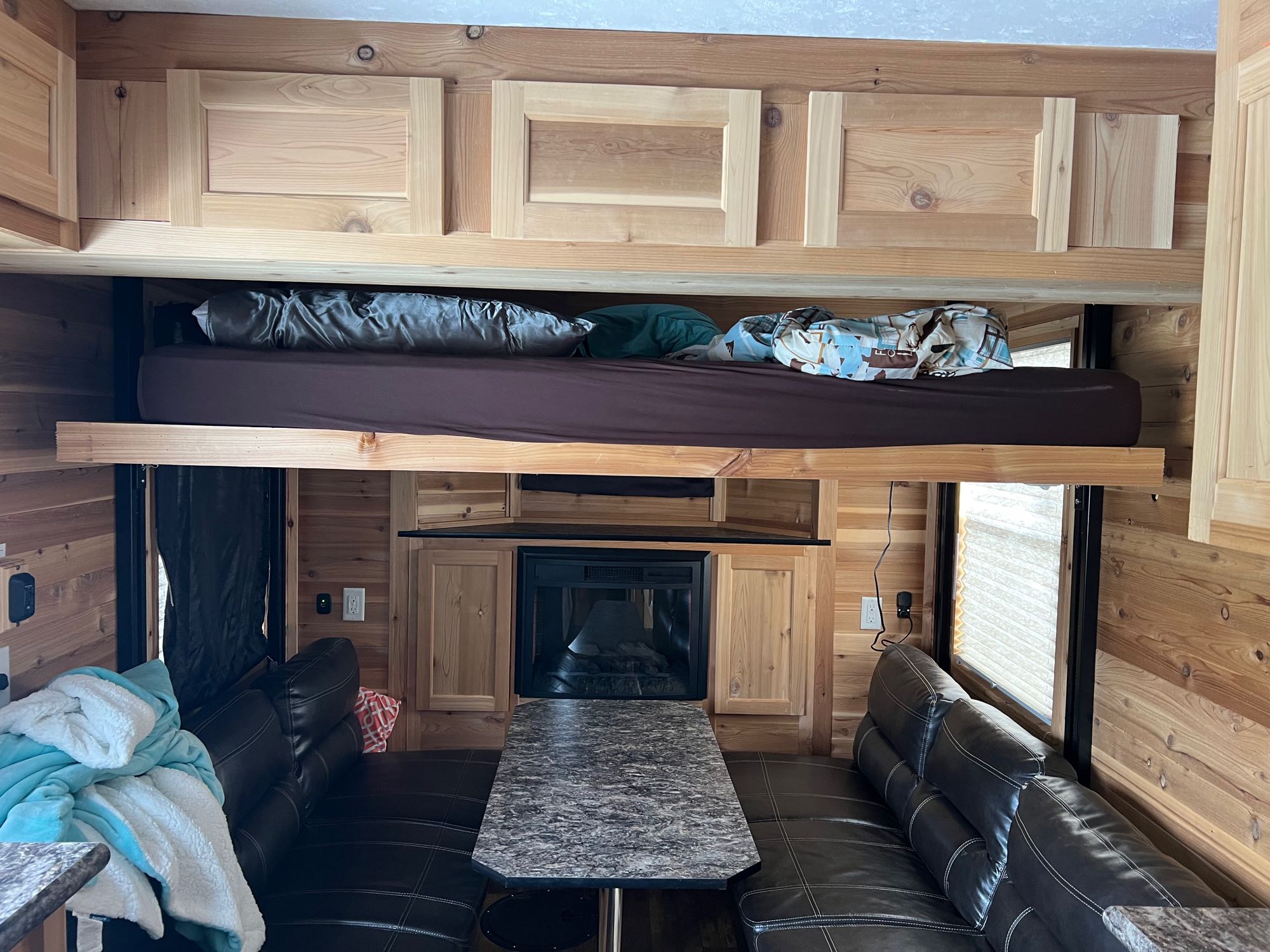 Cozy wood-paneled rv rental interior: upper bunk bed, lower seating with dinette table, and fireplace.. Ice Castle Fish Houses Ice Castle Fish Houses Trailer 2022