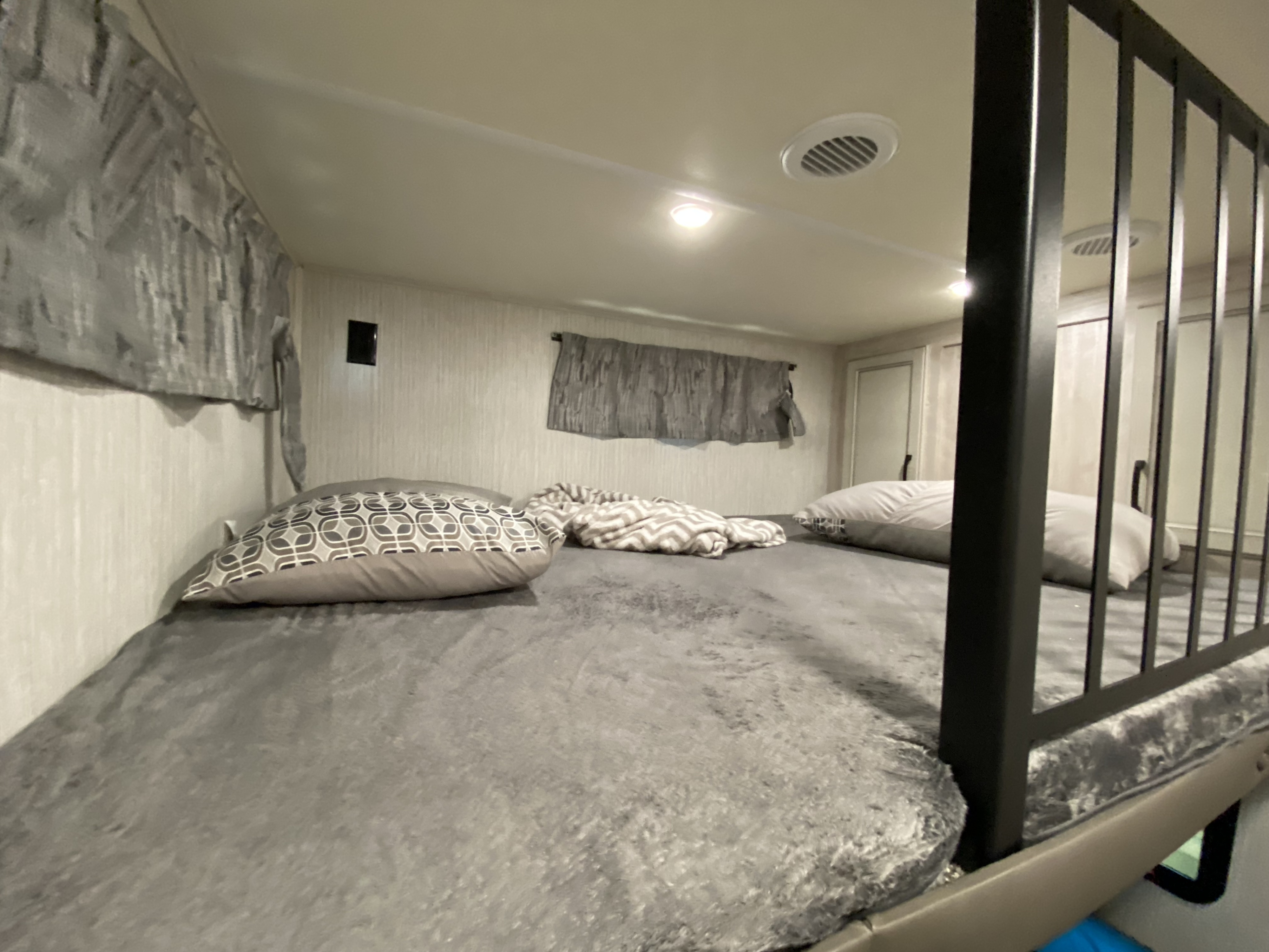 RV rental bunk bed: cozy grey bedding, decorative pillows, safety rail, and ceiling vent visible.. Forest River Sandpiper 2022