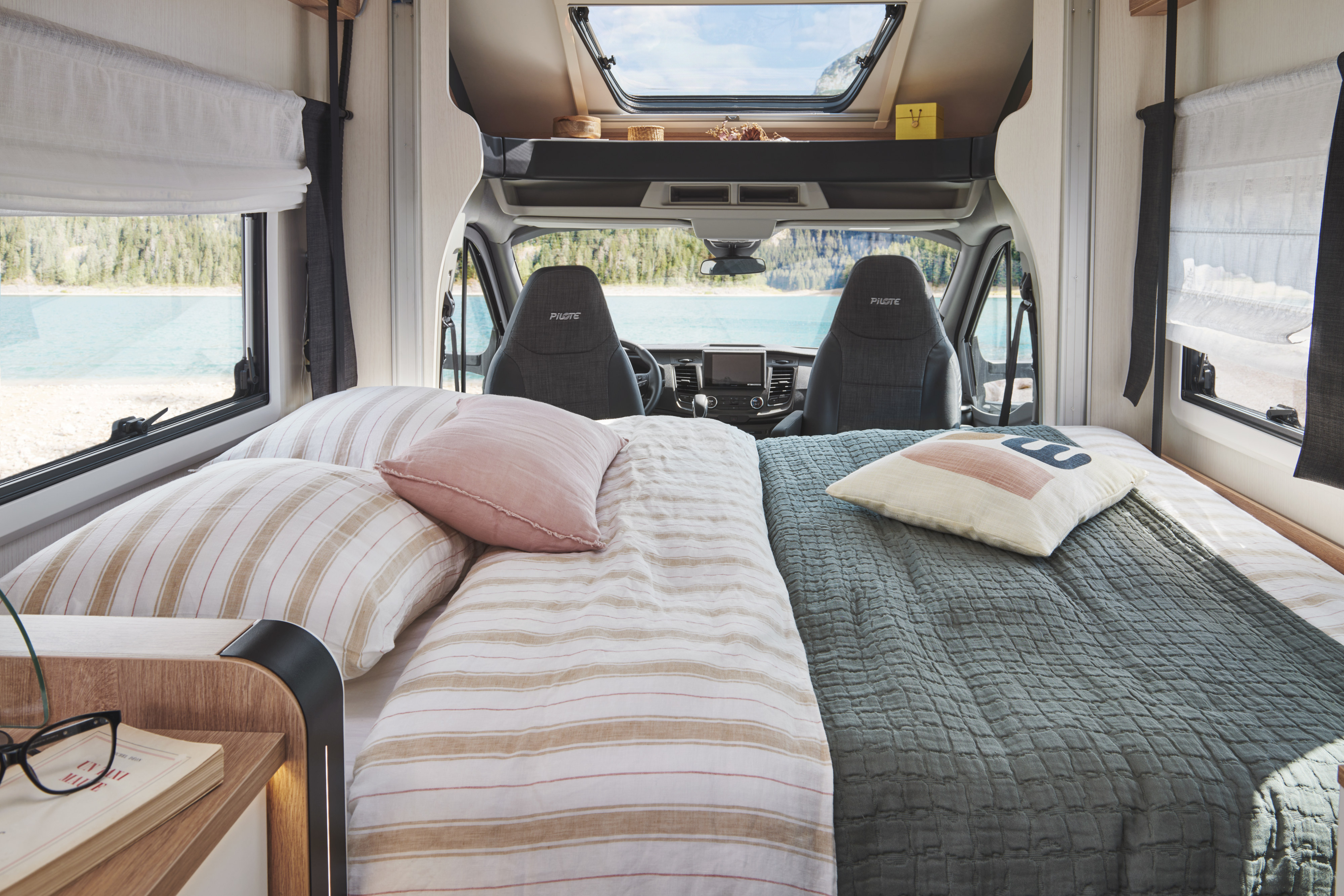 Plush RV rental bed with serene lake views; camper's front cabin and driver's seat visible, ready for adventure.. PILOTE A696G Atlas 2025
