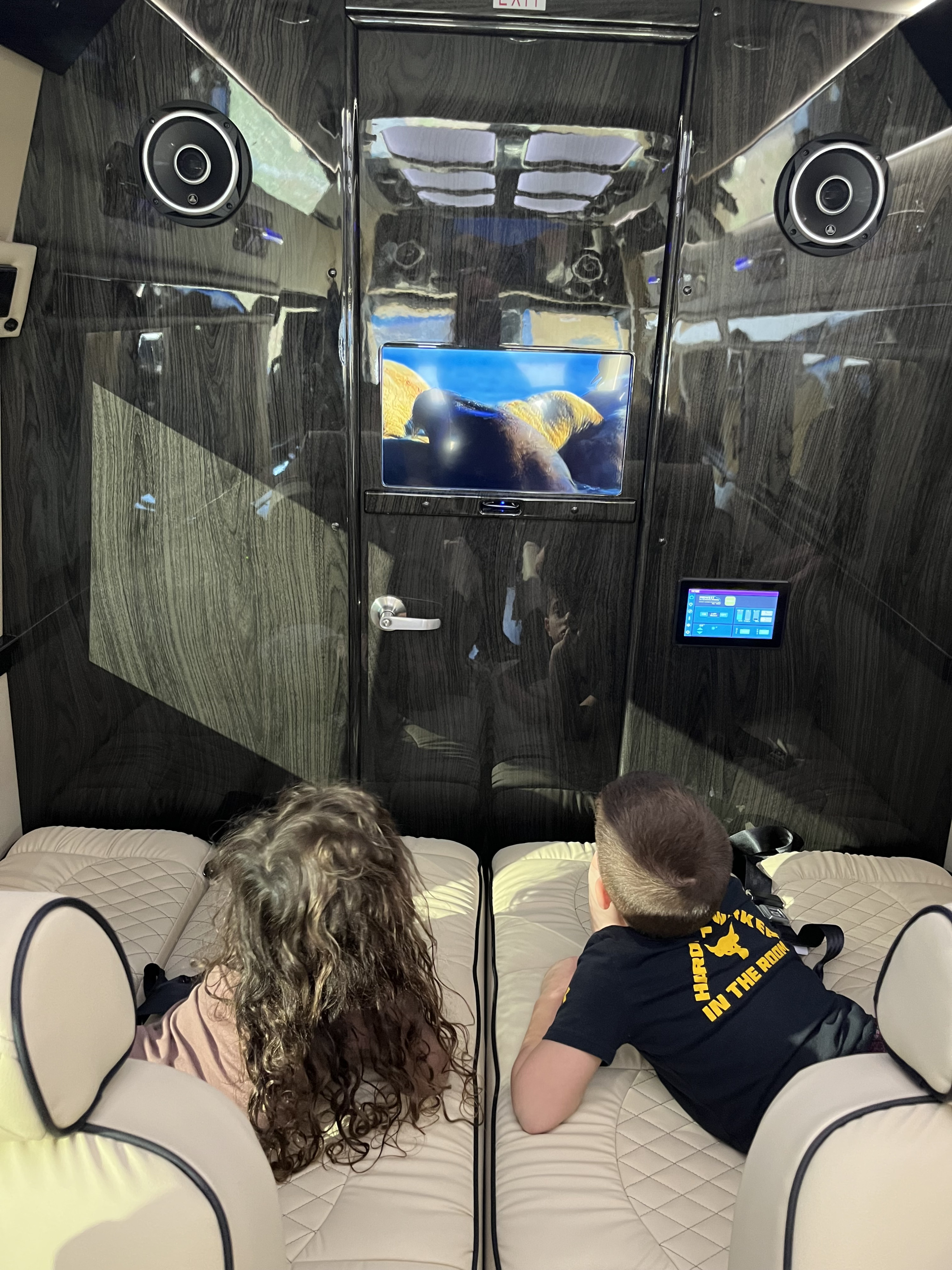 Kids enjoy a movie on the screen in a luxury RV rental, with comfortable seating and a sleek wood-grain interior.. Ultimate Toys Ultimate Coach 2024