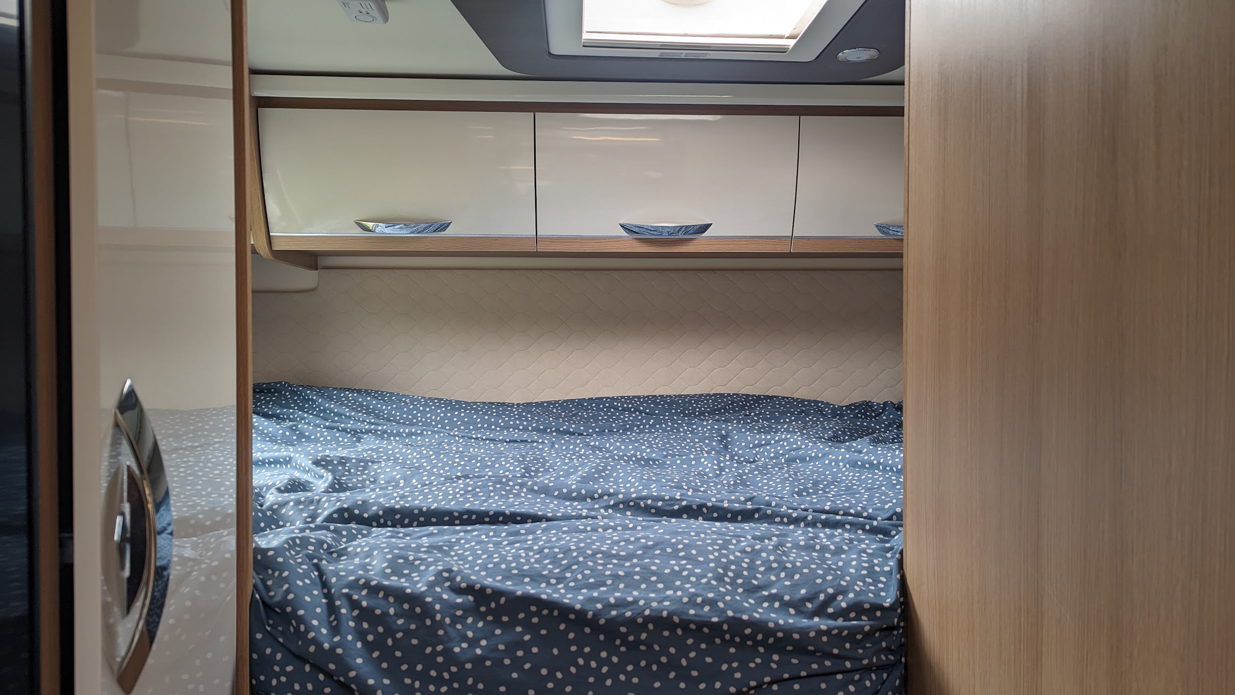 Inside an RV rental: a neat bedroom with a blue dotted duvet, ample overhead storage, and a skylight above the bed.. McLouis Fusion 360 2023