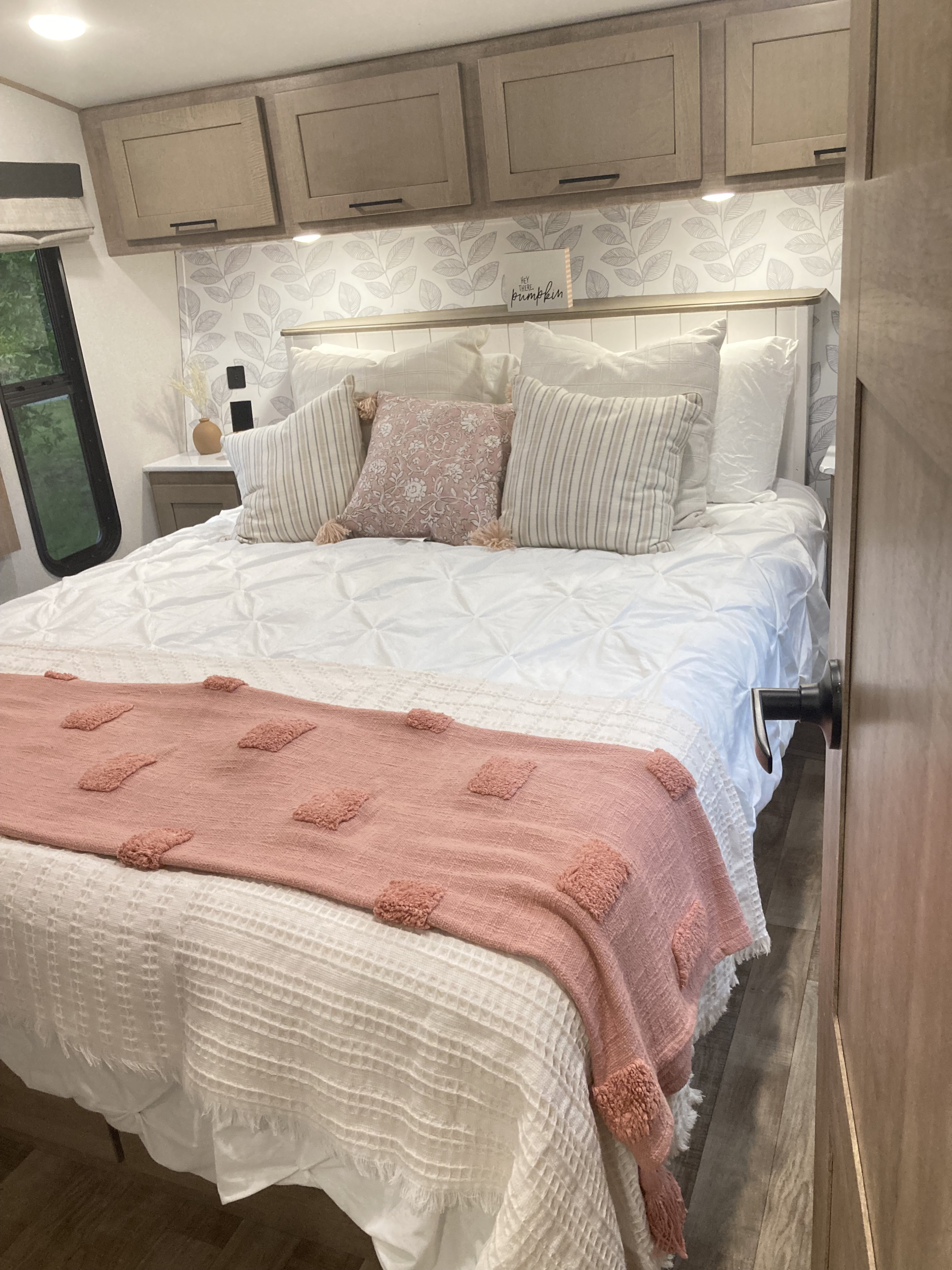 Cozy RV rental bedroom with white bedding, pink throw, decorative pillows, modern wood cabinets, and a view outside.. Keystone RV Arcadia 2022