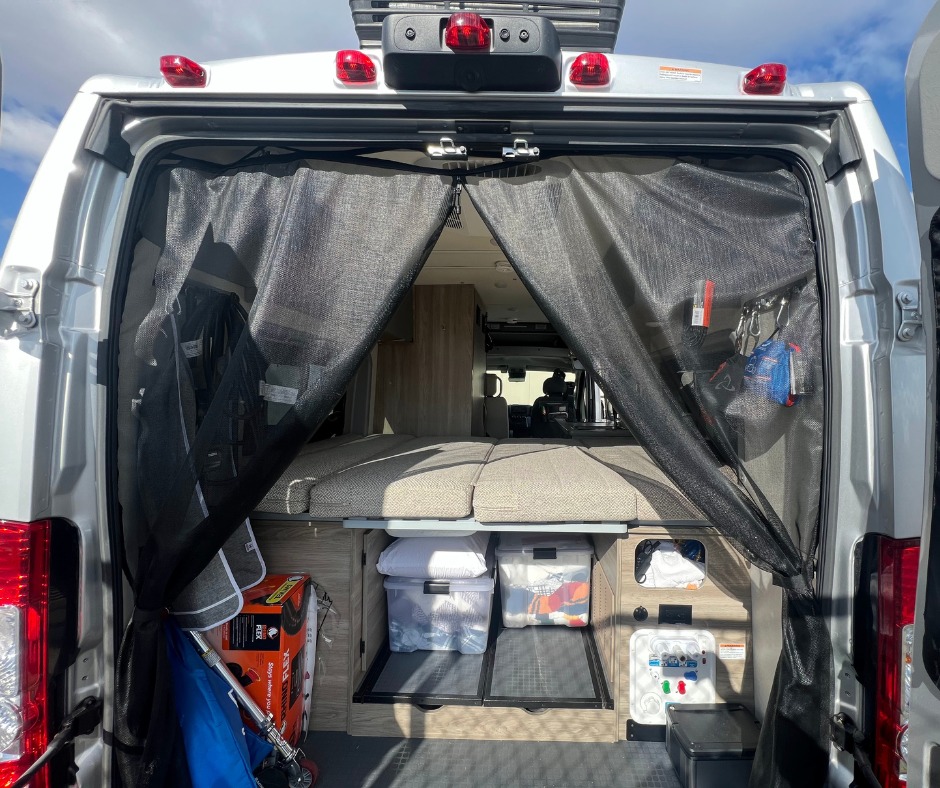 Inside an RV rental: rear view shows a mesh bug screen, modular bed, storage, and utility panel.. Winnebago 59PX 2021