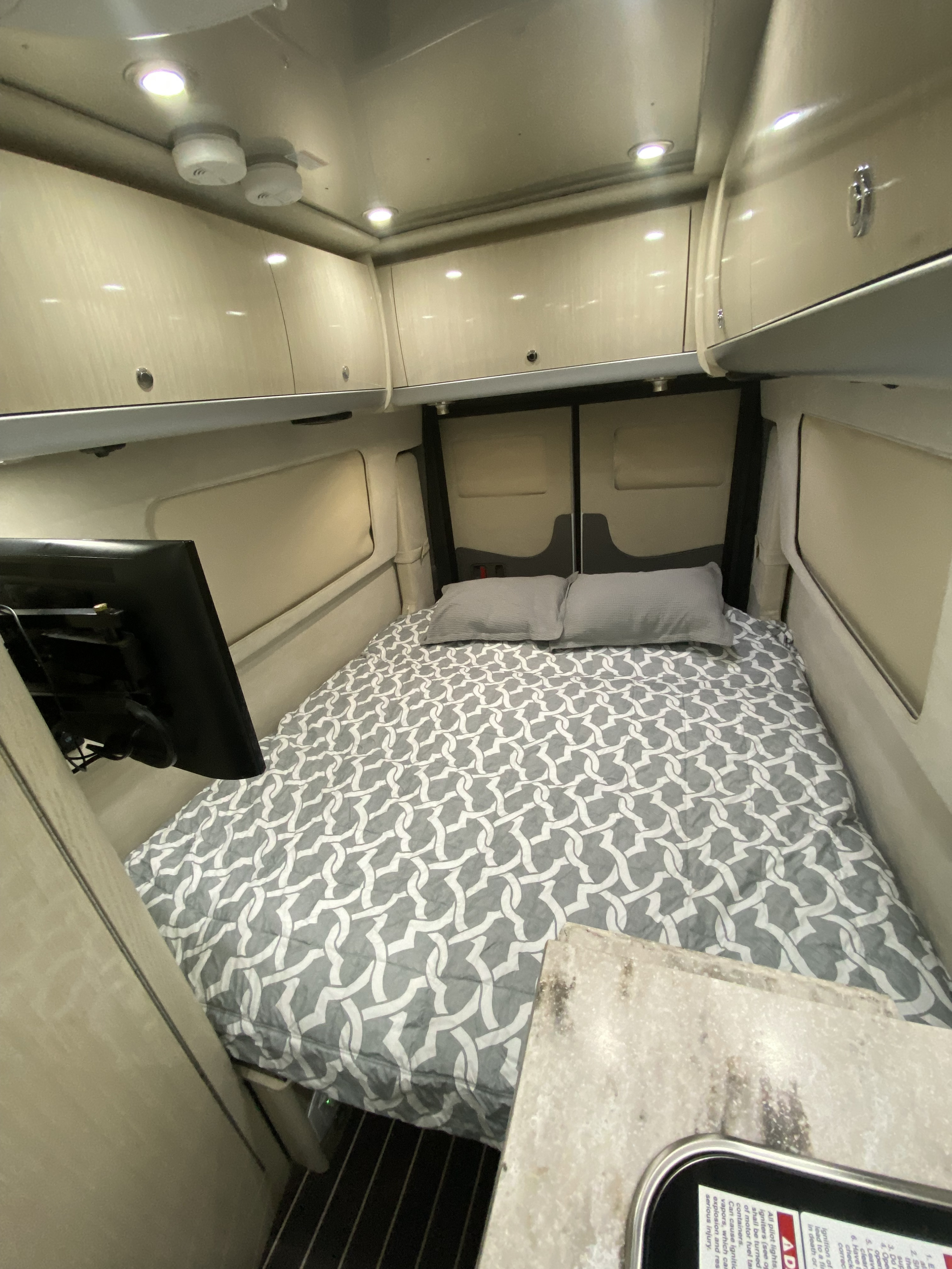 Cozy RV rental bedroom featuring a bed with patterned duvet, two pillows, overhead cabinets, and a TV.. Airstream Interstate 2017