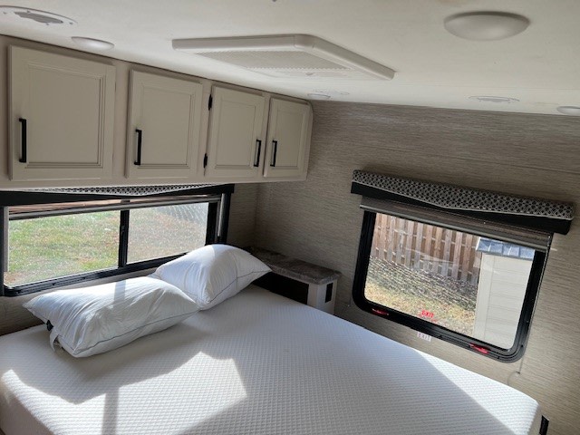 Step inside this cozy RV rental bedroom. Features a comfy bed, white pillows, overhead cabinets, and outdoor views.. Thor Chateau Victory 2022