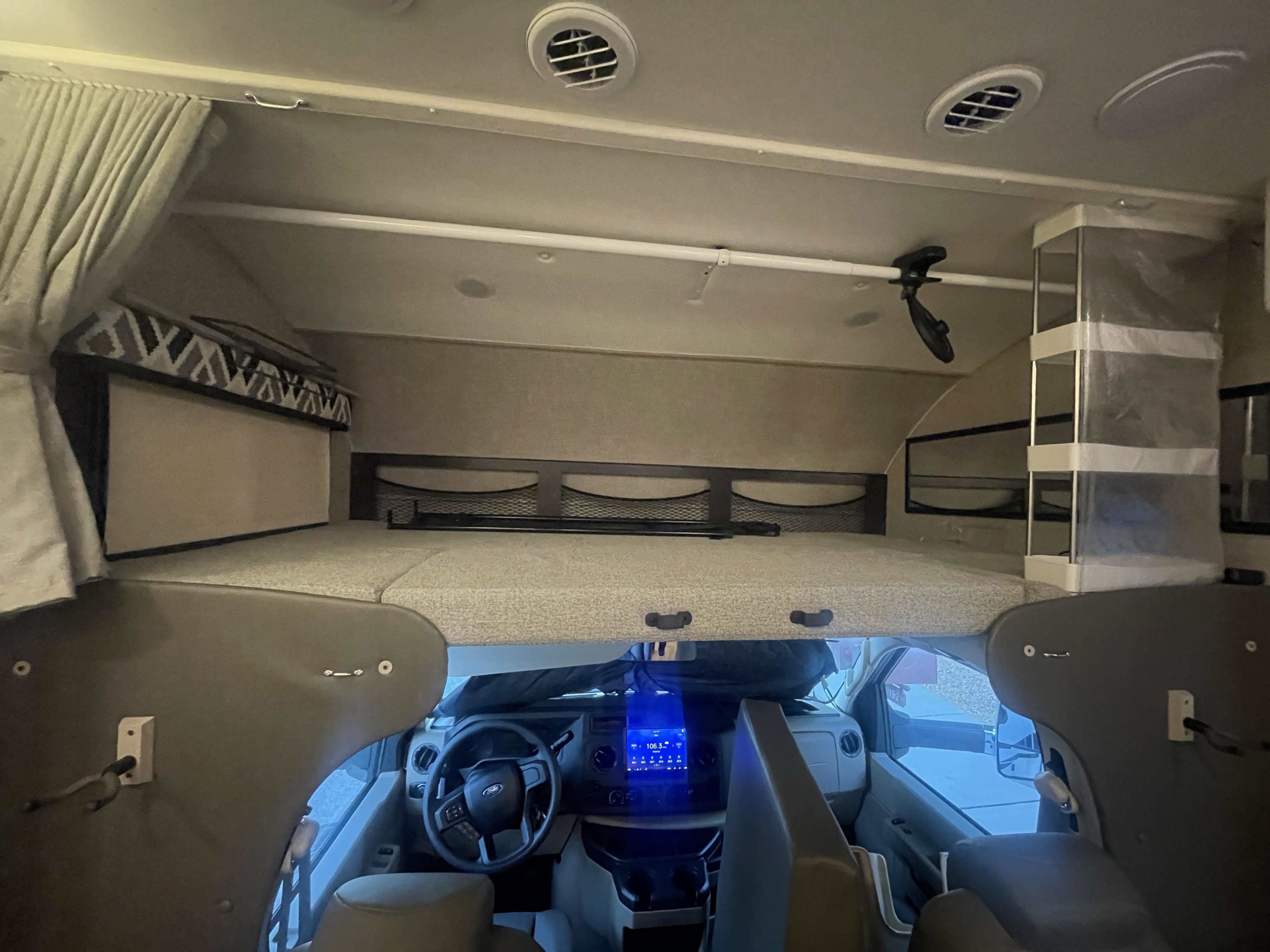 Inside an RV rental: Overhead bunk bed, privacy curtains, and the front cab with steering wheel and dashboard.. Jayco Greyhawk 2022
