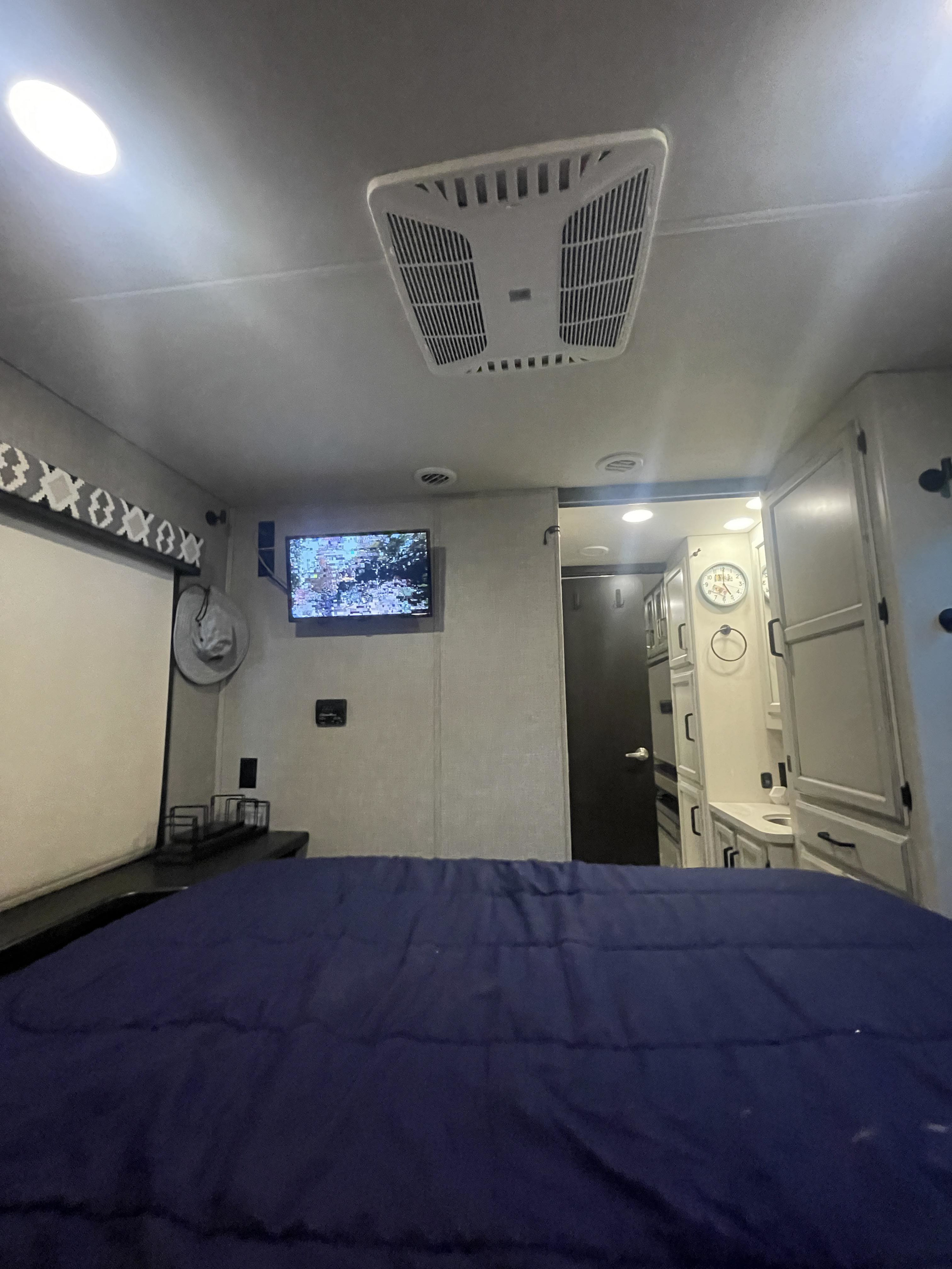 RV rental bedroom: dark blue bed, wall-mounted TV with static, AC unit. Bathroom with clock visible through doorway.. Jayco Greyhawk 2022