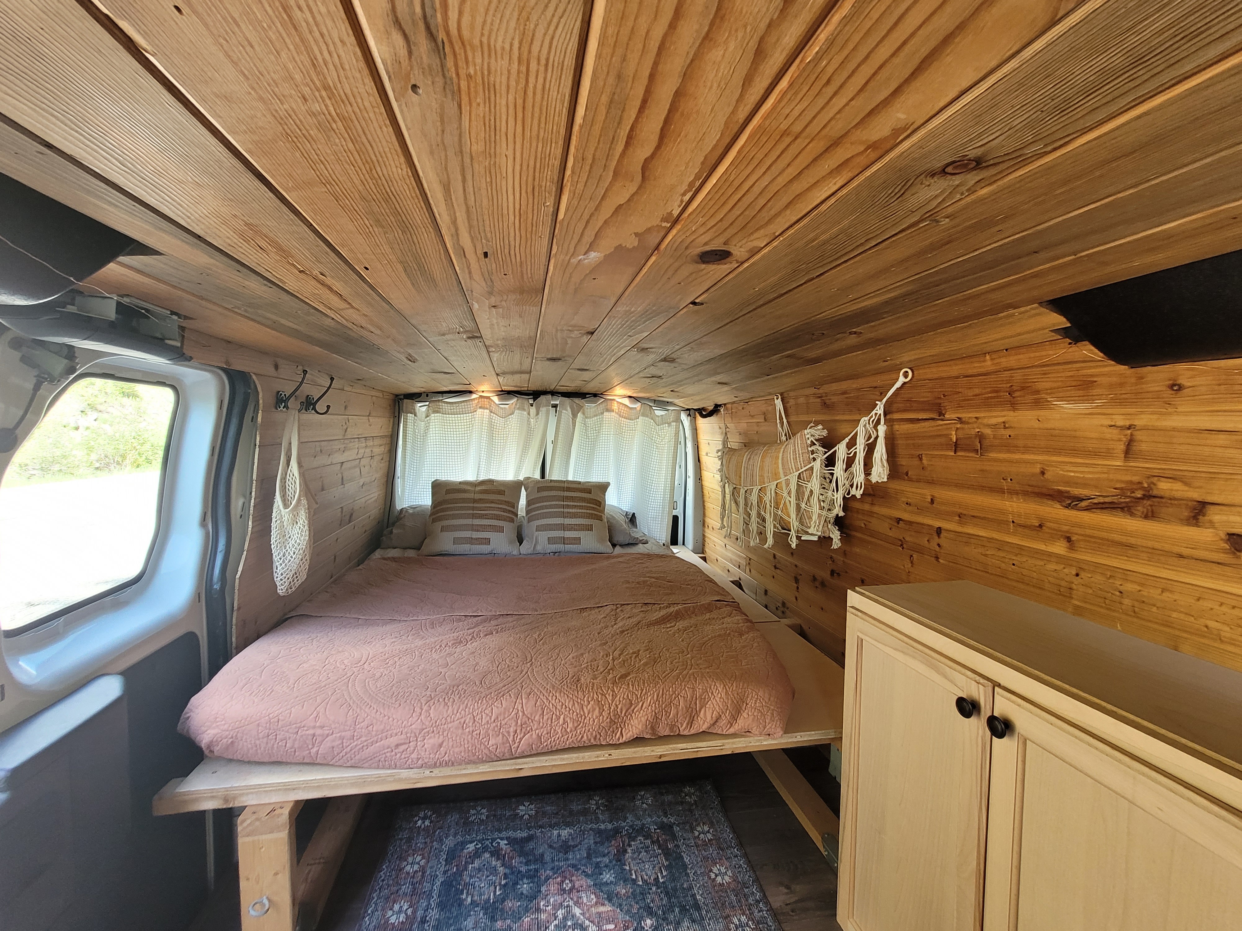 Cozy, wood-paneled RV rental interior: bed with pillows & comforter, hanging storage, and cabinet. Perfect for van life.. Ford Transit 2016