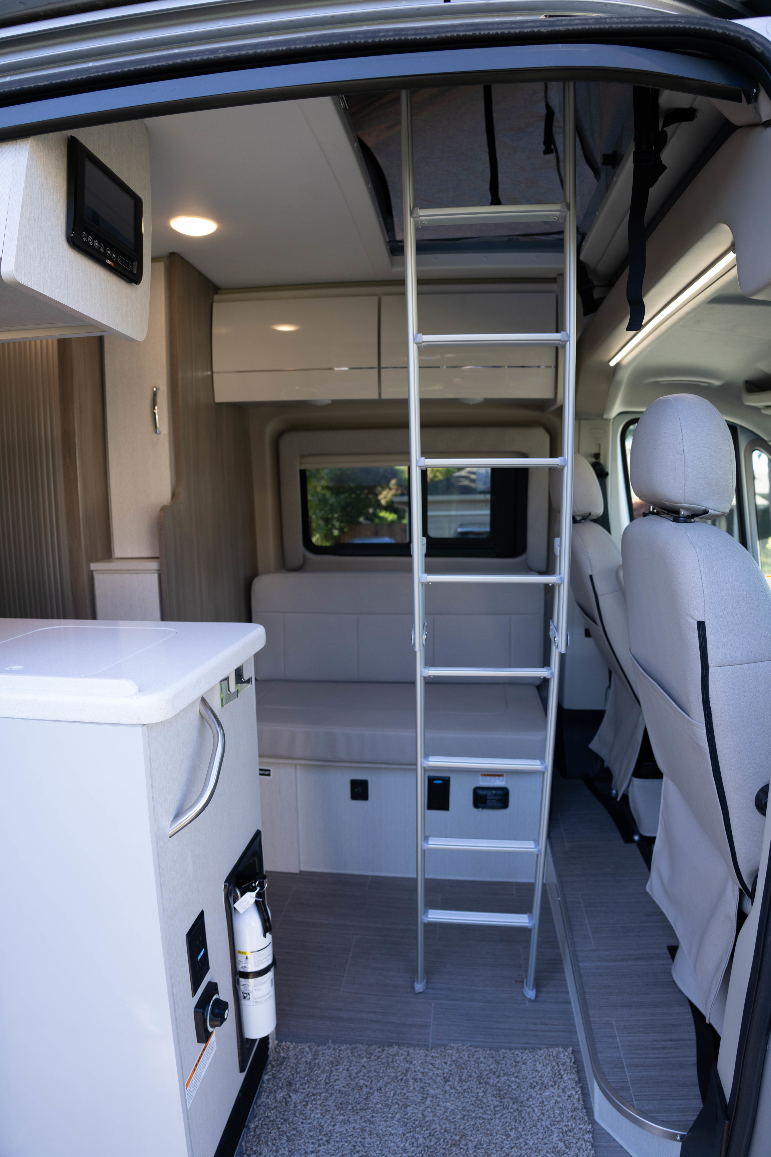 Inside an RV rental: a ladder to the upper bed, compact kitchen with fire extinguisher, rear seating, and driver's seats.. Thor Tellaro 2023
