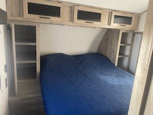 Experience comfort in this RV rental bedroom featuring a blue-covered bed, overhead cabinets, and side storage shelves.. Forest River Cherokee Alpha Wolf 2021