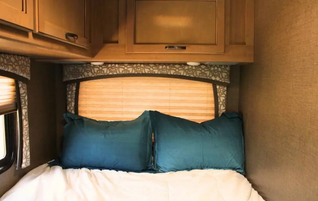 Cozy RV rental bedroom with teal pillows on a bed, window, and overhead cabinets. Compact comfort for travel.. Thor Chateau 2018