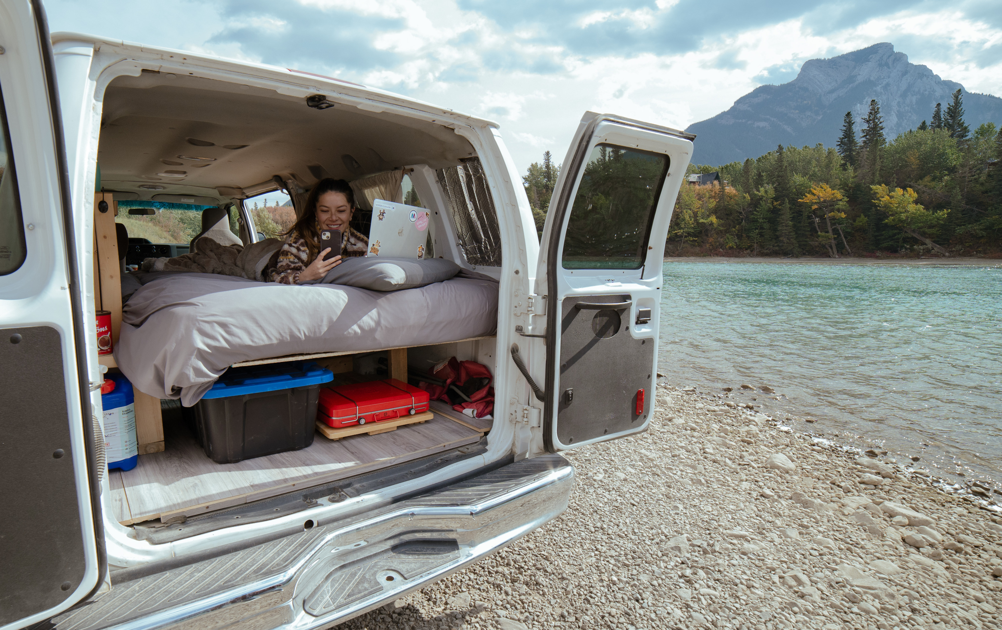 RV rental comfort: Woman works & relaxes in a camper van with doors open to a stunning river & mountain view.. Glacier Campers GlamperVan 2014