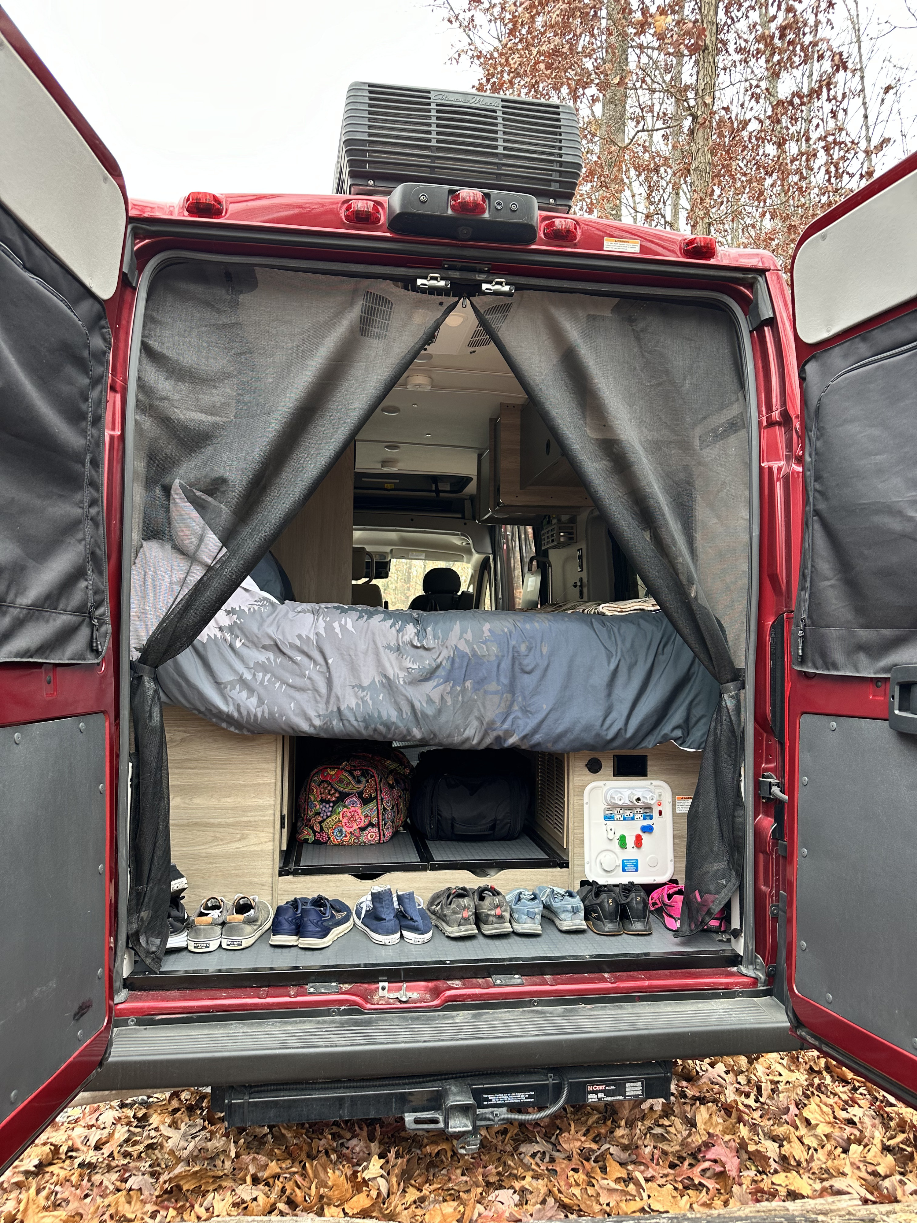 Red RV rental's rear doors open, revealing a cozy bed, shoe storage, and outdoor hookups amidst autumn leaves.. Winnebago Solis 2022