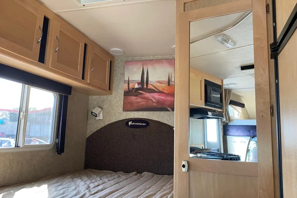 Inside an RV rental: a comfortable bedroom area with overhead storage, a window, and a reflection of the compact kitchen.. Adventurer Adventurer Motorhome 2010